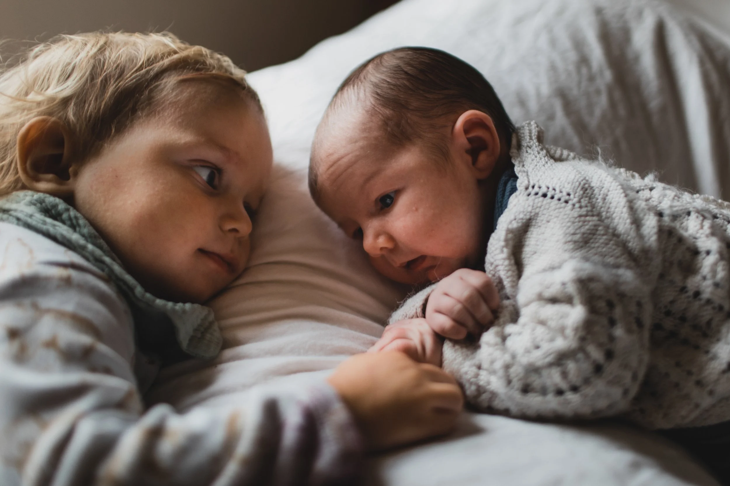 A young girl and a baby lying on a bed, facing each other, holding hands and looking into each other's eyes.