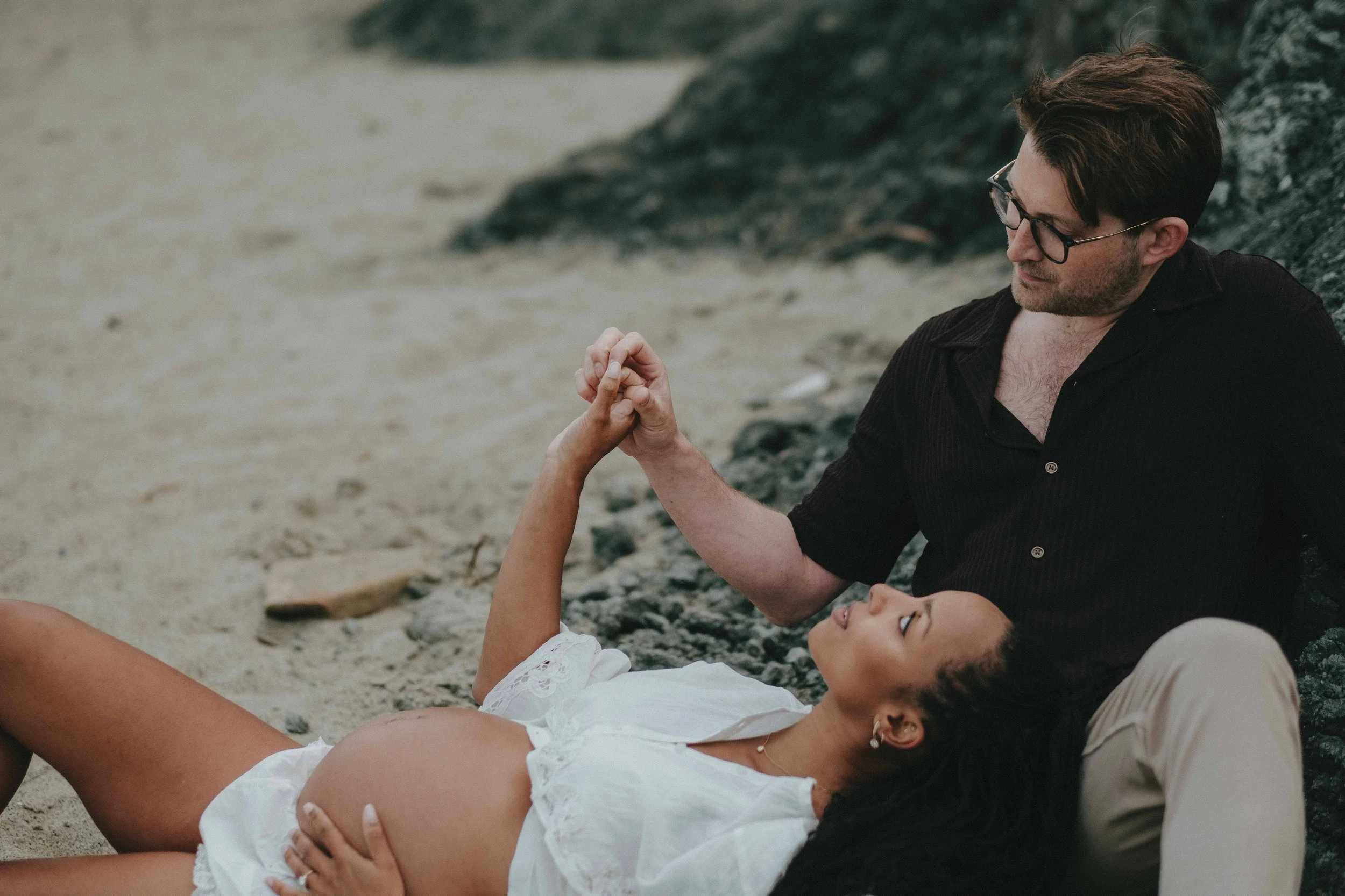A pregnant woman lying on sand at the beach, holding hands with a man sitting next to her, both smiling and gazing at each other.