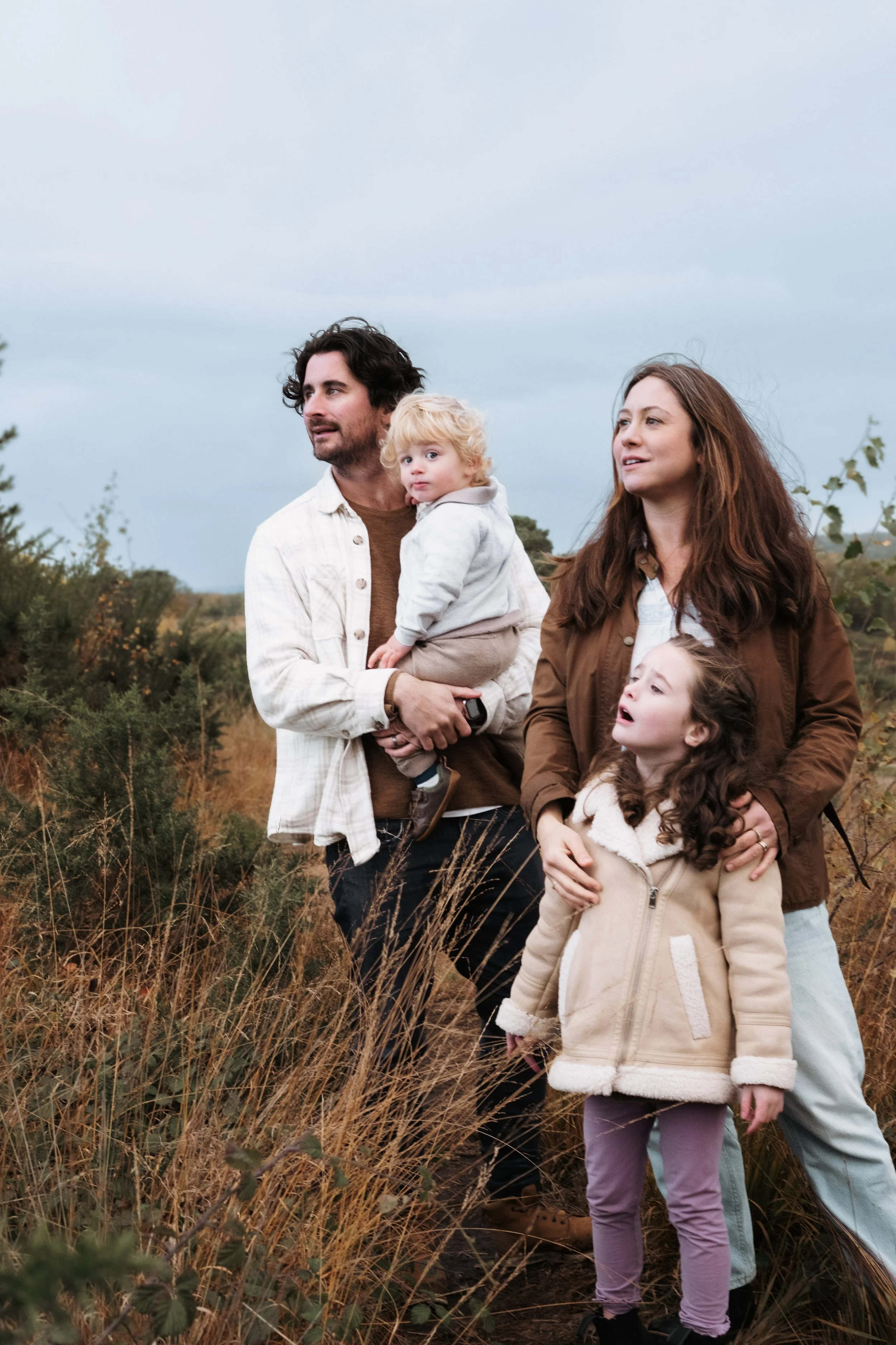A family of four standing outdoors in a field with tall grass and bushes, under a cloudy sky.