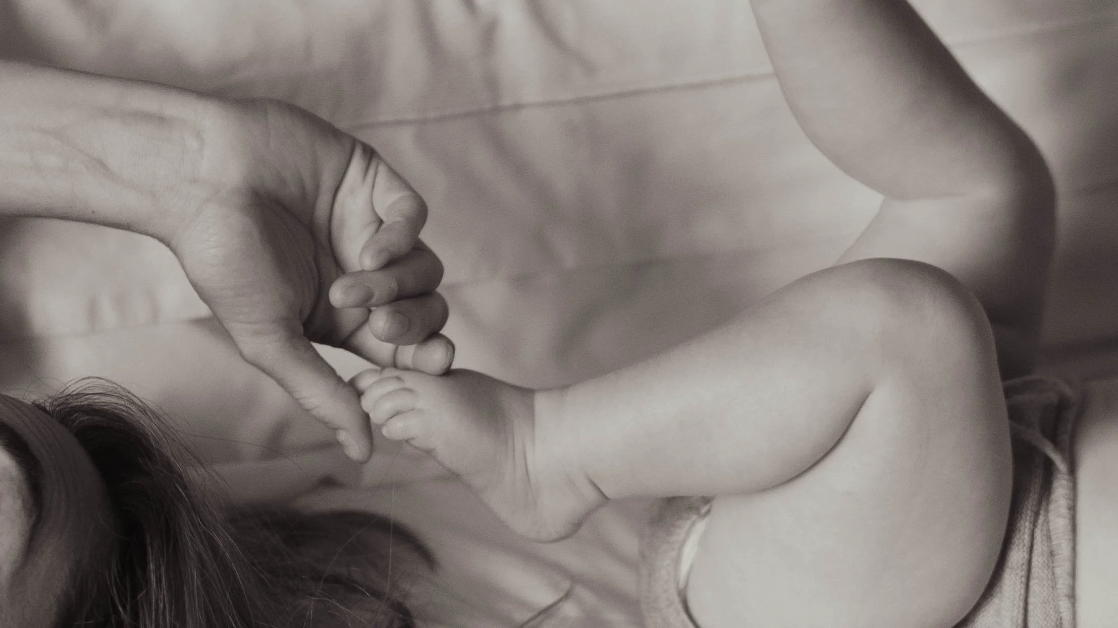 A person's hand gently holding a baby's foot, with the baby's leg visible and partly covered by a fabric, in a black-and-white photograph.