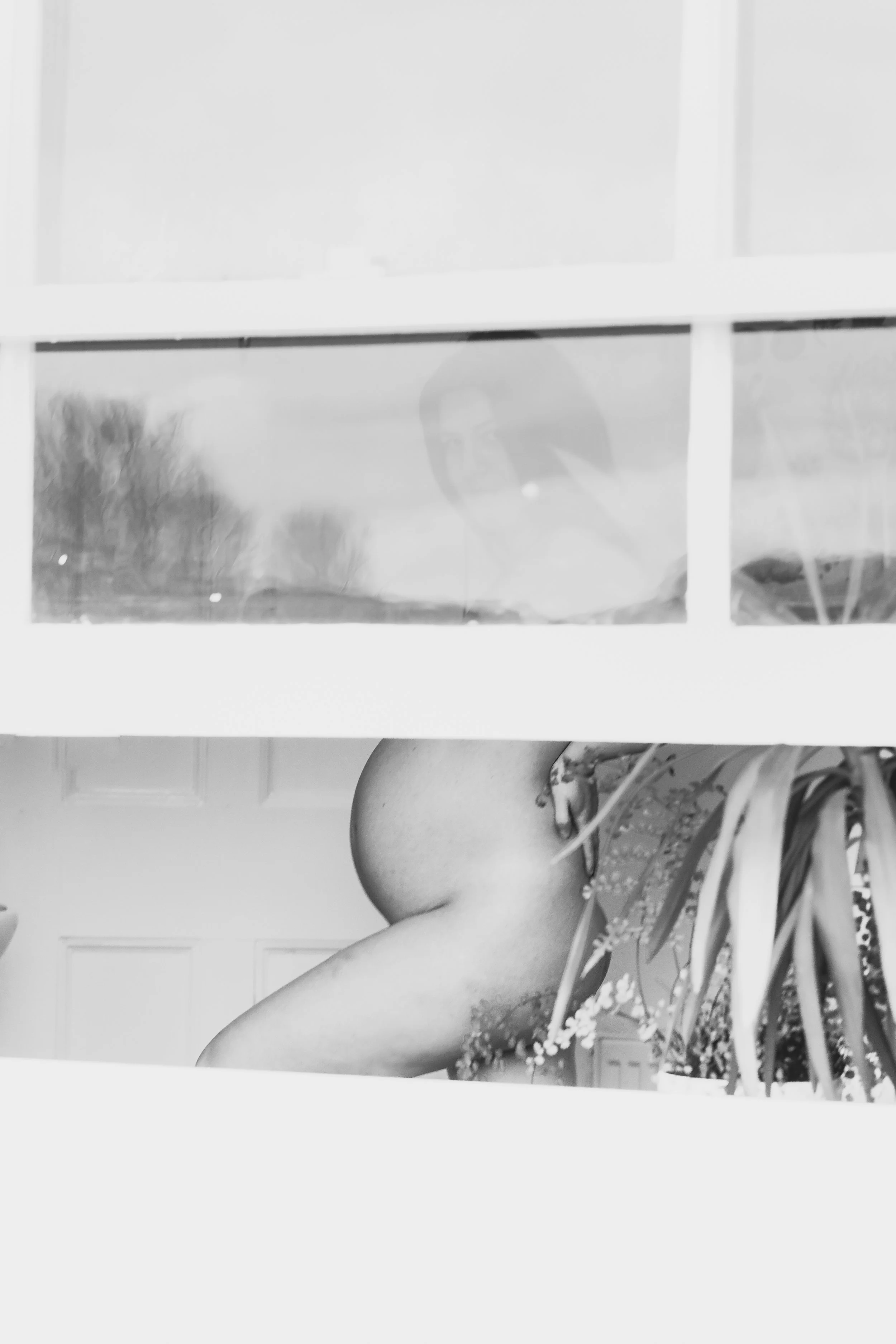 A black and white photo showing a person with long dark hair, viewed through a glass window, with the reflection of trees outside. The lower part of the image shows a person's bare leg resting on their knee in a room with a door in the background, an