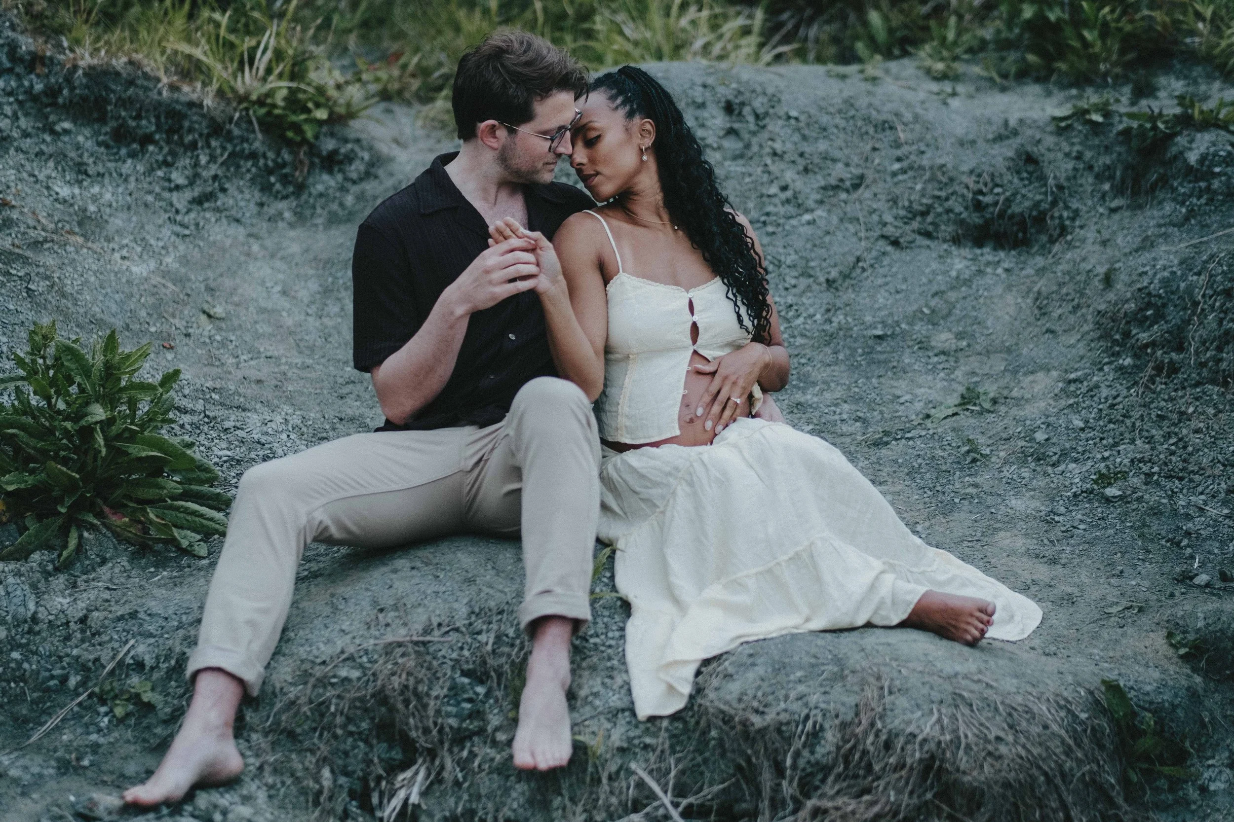 A couple sitting outdoors on a rocky surface, embracing each other closely. The man is wearing glasses, a black shirt, and light-colored pants. The woman, with long curly hair, is dressed in a cream-colored dress and shows her pregnant belly. They ar