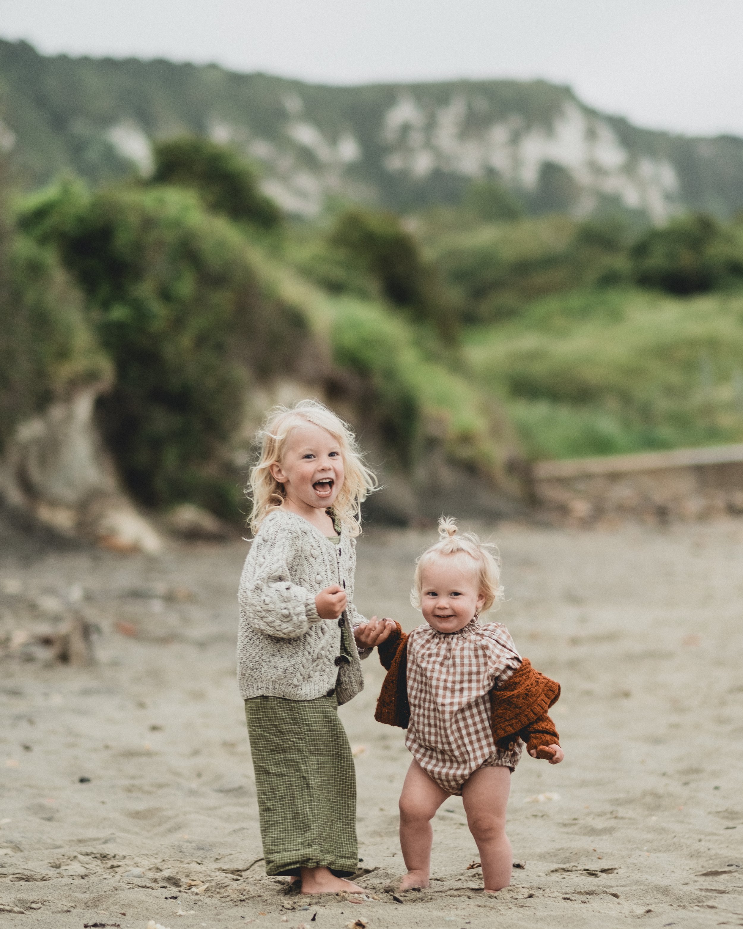 Two young children, a girl with blonde hair and a boy with blonde hair in a top knot, are standing barefoot on a sandy beach, smiling and holding hands. Behind them are green hills and a cloudy sky.