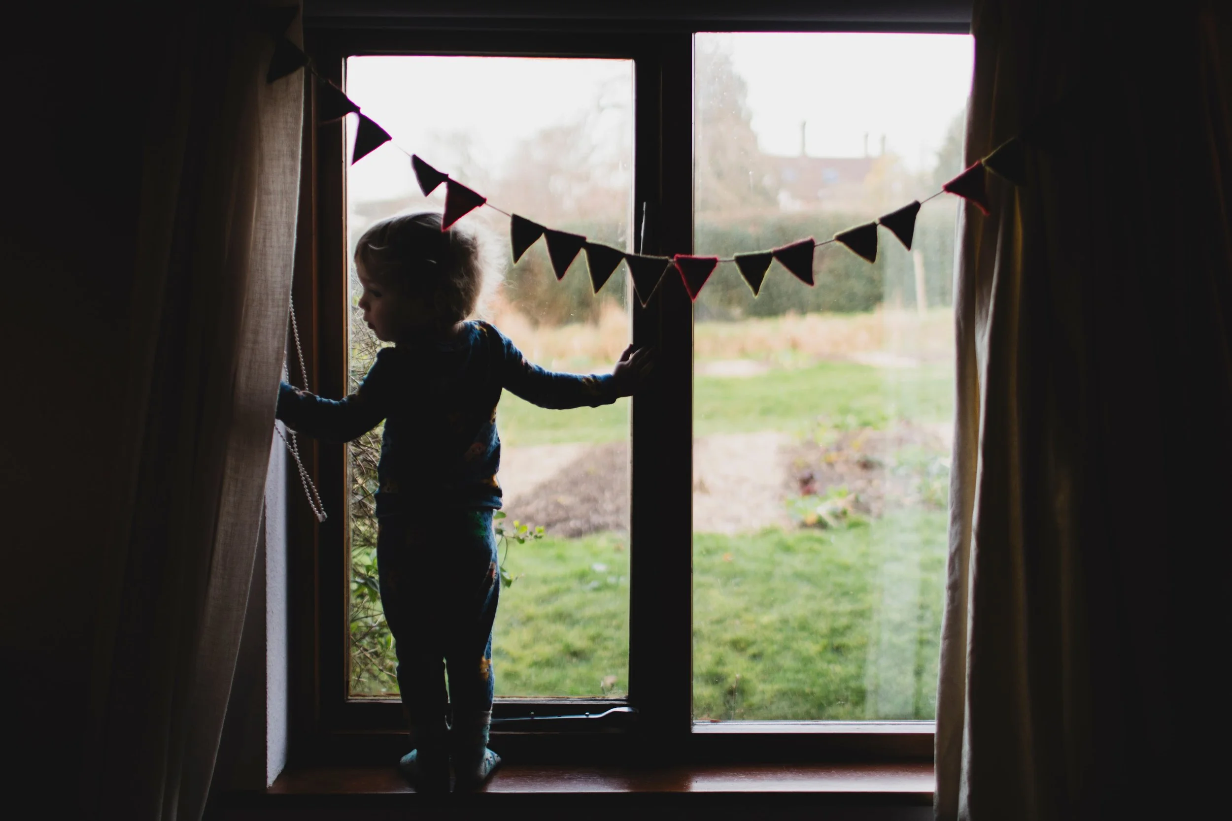 Child standing on a window sill, looking outside, with curtains pulled back on both sides and a string of triangular flags hanging across the window.