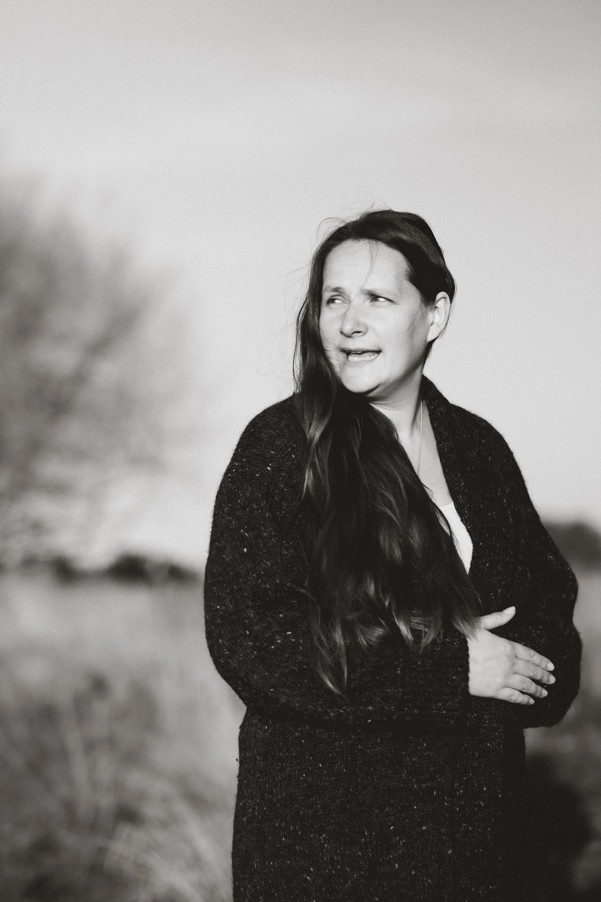 A woman with long dark hair, wearing a black textured sweater, standing outdoors with her arms crossed, looking to the side with a slight smile, over a blurred natural background.