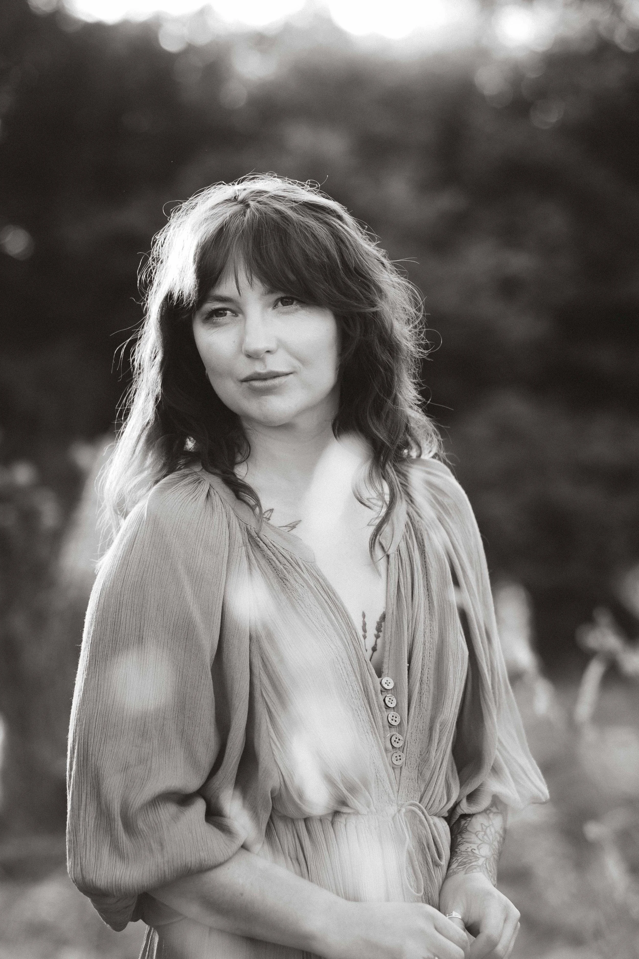 Black and white photo of a woman outdoors with trees in the background, looking slightly to her left with a soft smile.