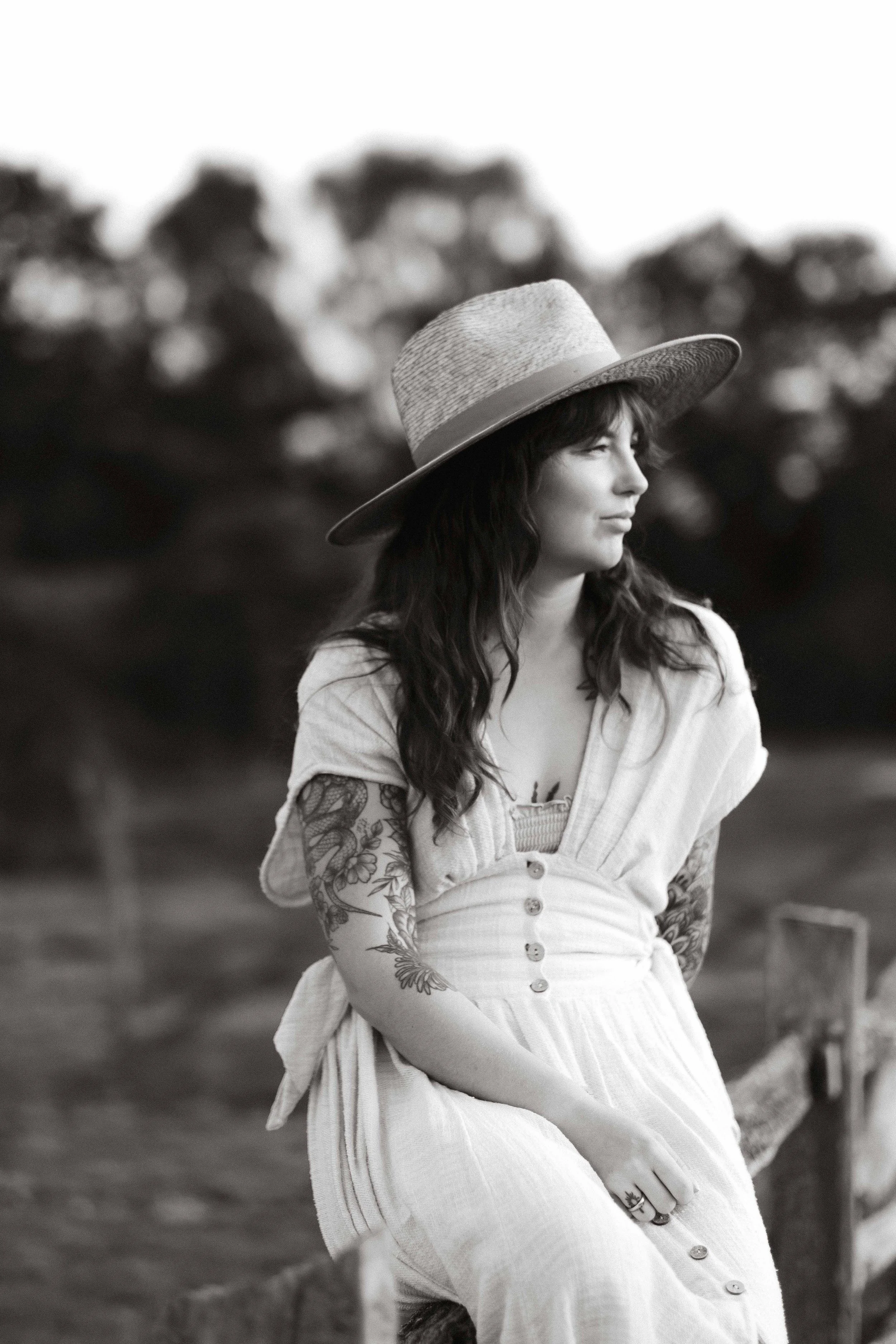 A woman with long dark hair and tattoos on her arm, wearing a wide-brimmed straw hat, a buttoned dress, and a ring, sitting outdoors by a wooden fence with trees in the blurred background, in black and white.