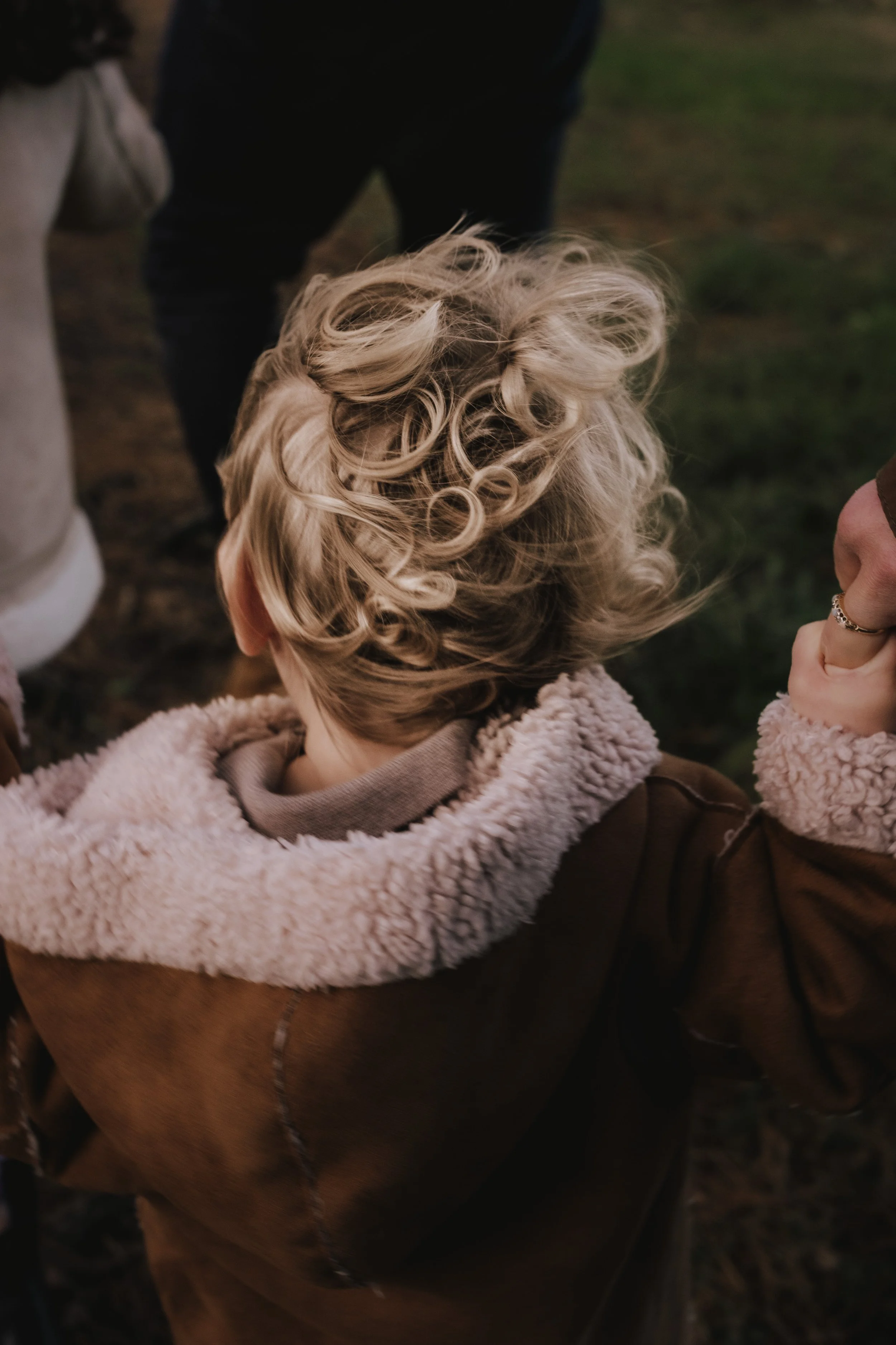 Child with curly blonde hair wearing a brown coat with a sherpa-lined hood, being held by an adult outside.