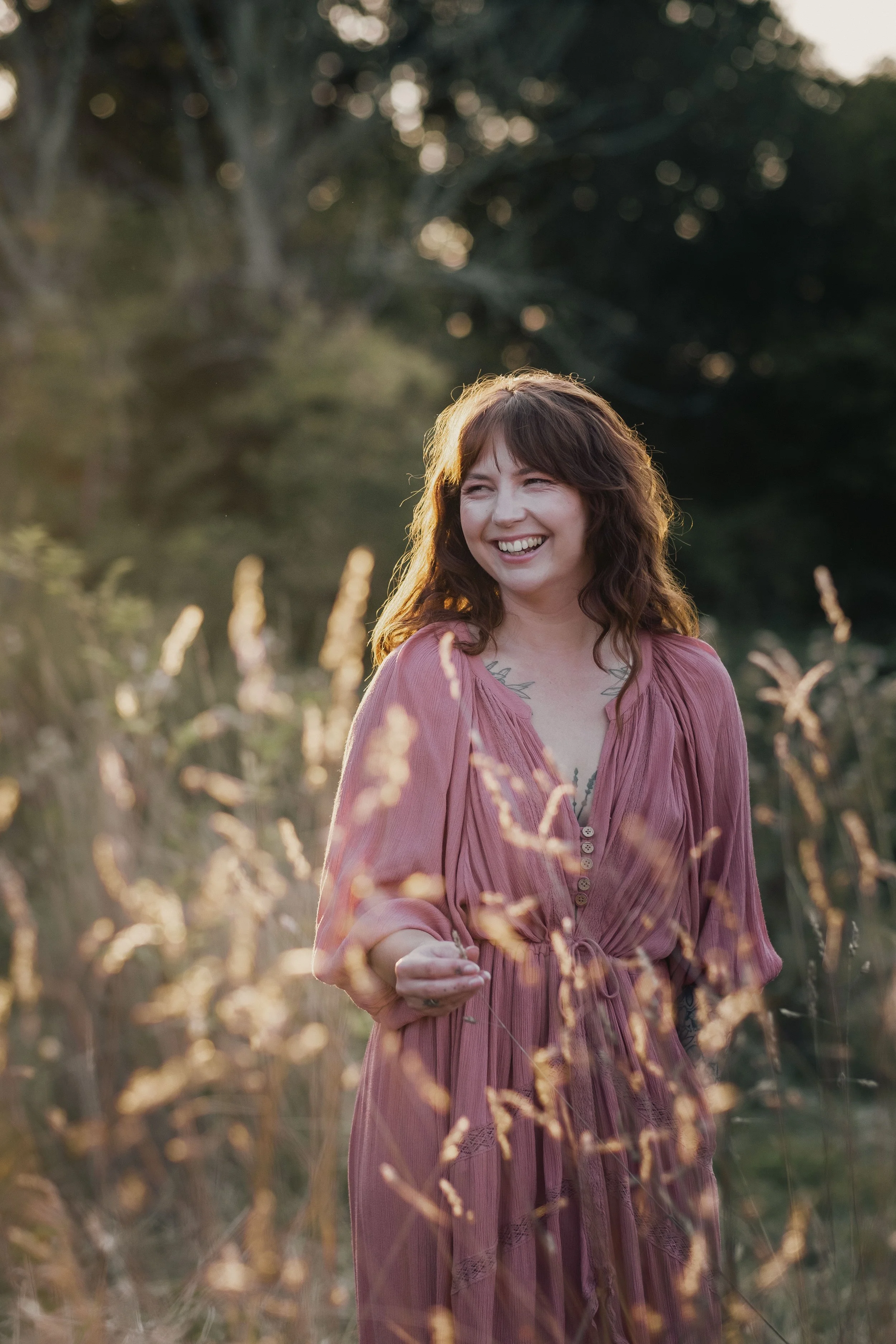 A woman with wavy brown hair, wearing a pink dress, smiling while walking through a field of tall grasses during sunset.