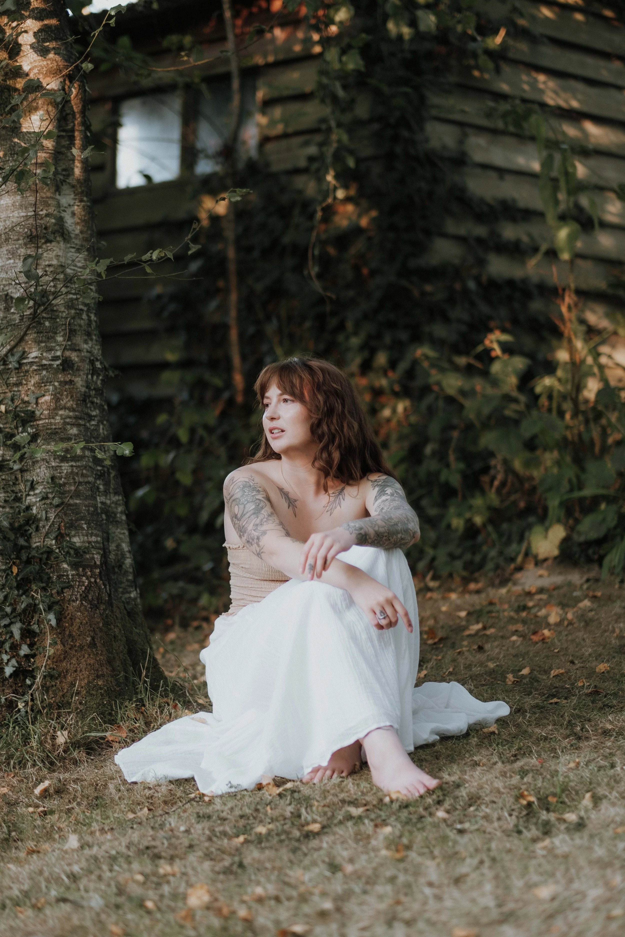 A woman with brown, curly hair, tattoos on her arms, sitting on the grass near a tree in a natural outdoor setting during daytime, with a wooden structure and green foliage in the background.