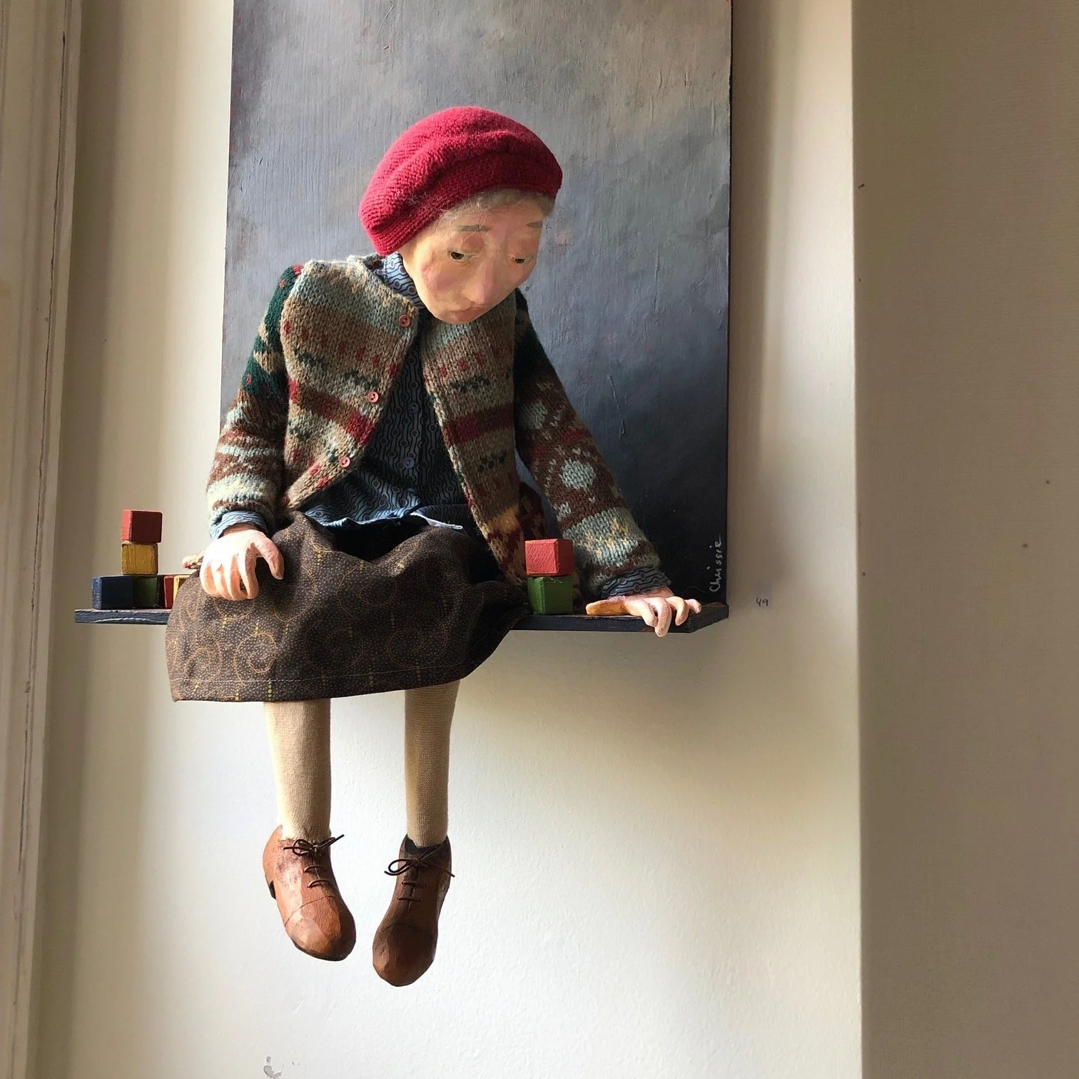 En konstnärlig skulptur av en äldre kvinna som sitter på ett svart bord, med en tygkappa, klänning, strumpor och skor, och en röd mössa, avbildad på en vägg.