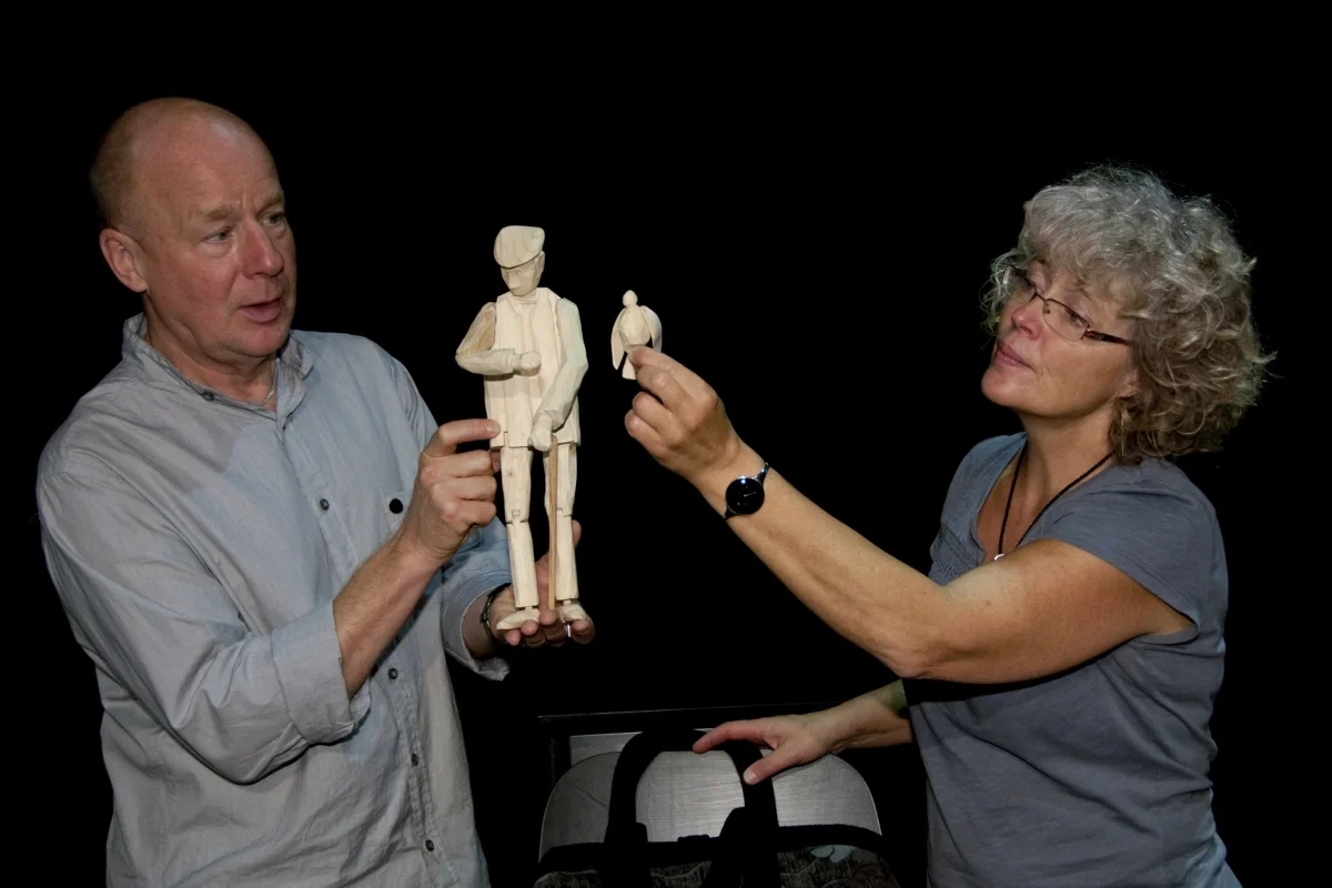 Två personer, en man och en kvinna, är under en presentation av en trädocka. Mannen håller dockan, en stiliserad docka av träklossar, och kvinnan pekar mot den.