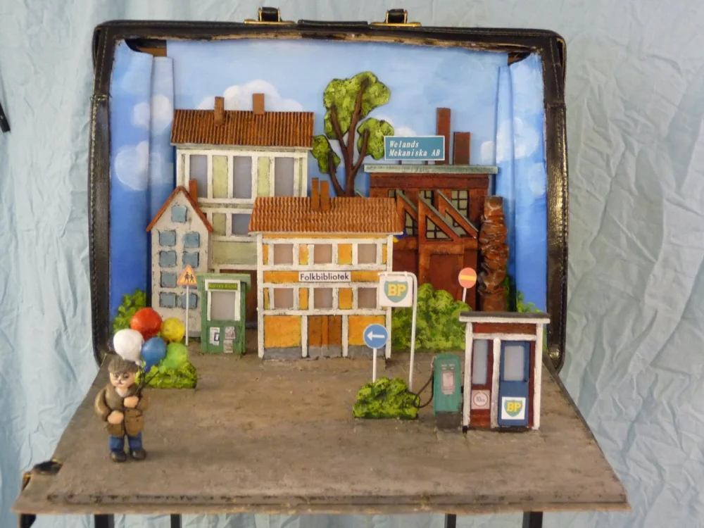En miniatyrstad med hus, en personfigur med ballonger, och olika skyltar och byggnader, inklusive ett bibliotek och en bensinstation, inuti en koffert.