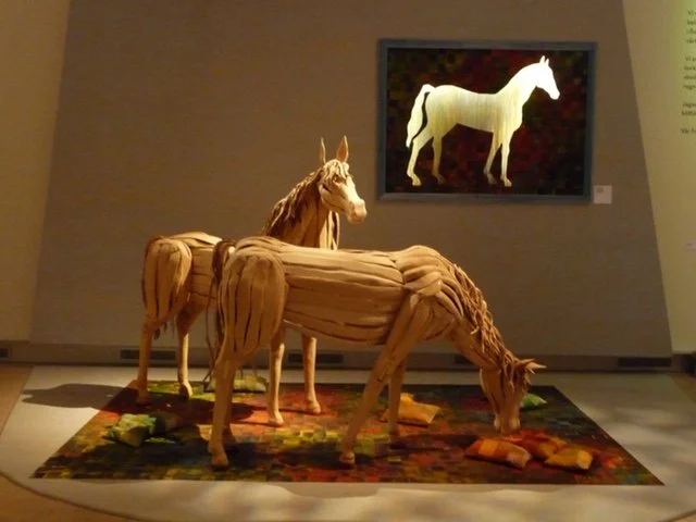 Skulptur av tre hästar på en matta framför en konstnärlig målning av en vit häst på en vägg.