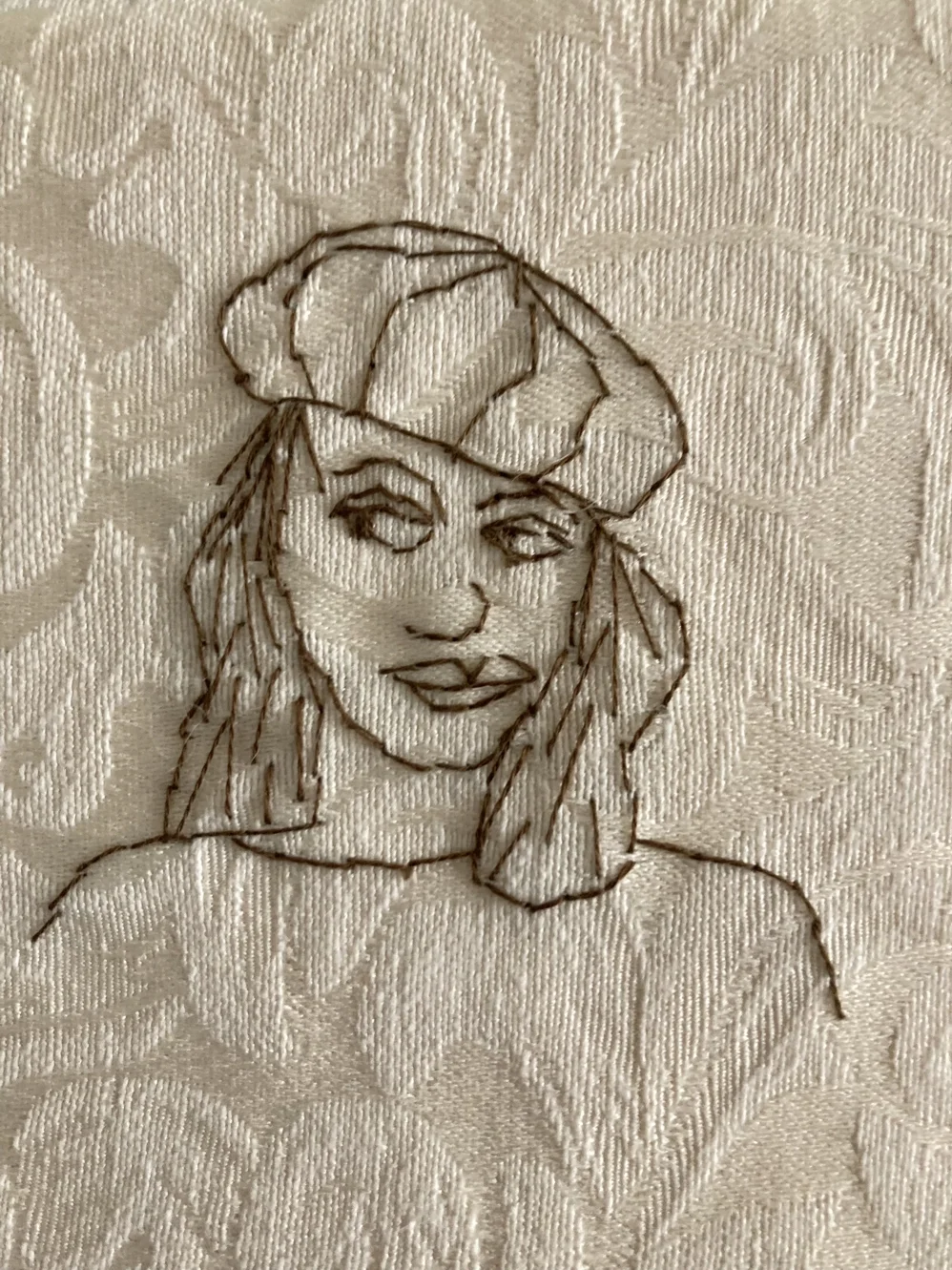 Mönsterbroderi av en kvinna med mössa och långa ögonfransar på ett beige tyg.