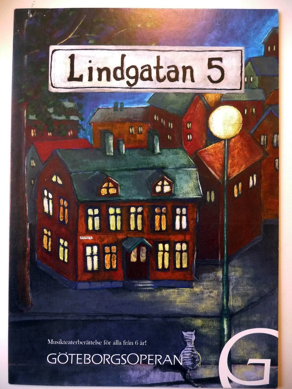 Festskrift för Göteborgsoperan med en tecknad landskapsbild av en stad med flerfärgade hus, ett gatuljus, och en katt som sitter på trottoaren.
