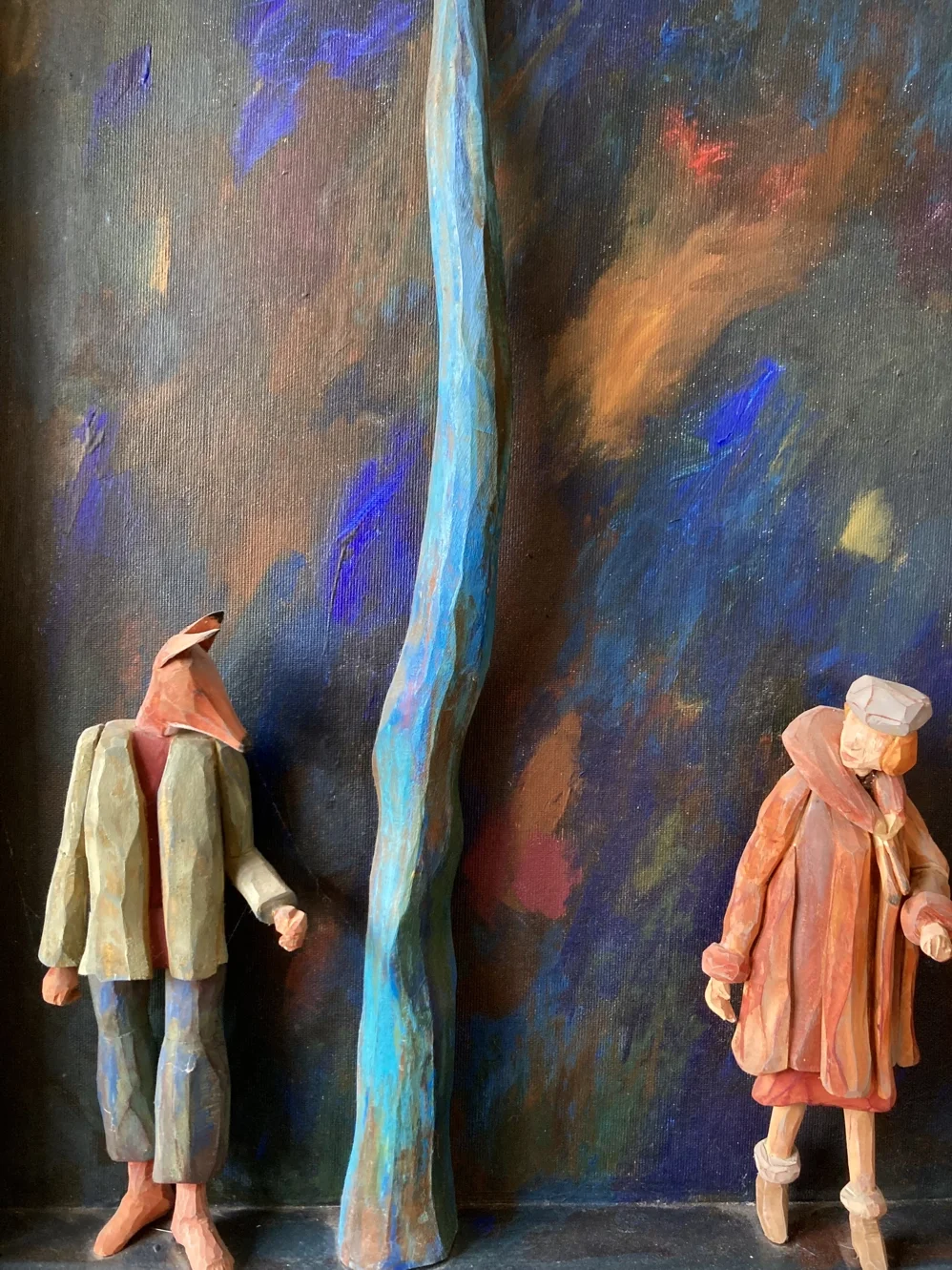 Två träfigurer av en man och en kvinna står framför en färgglad, abstrakt målarduk.