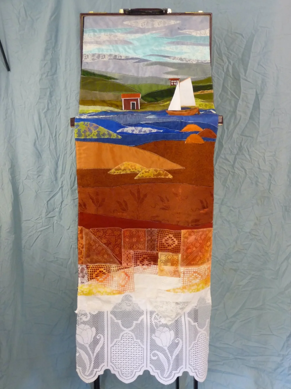 Textilt konstverk föreställande en landskapsbild med hav, hus och fyr, uppdelad i flera avsnitt med olika texturmönster och färger.