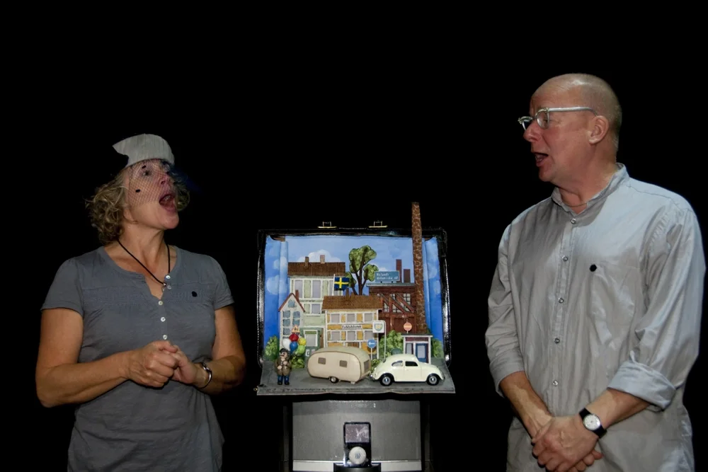 Två personer framför en låda med en miniatyrstad, en kvinna i grå t-shirt och hatt och en man i ljus skjorta, båda pratar.