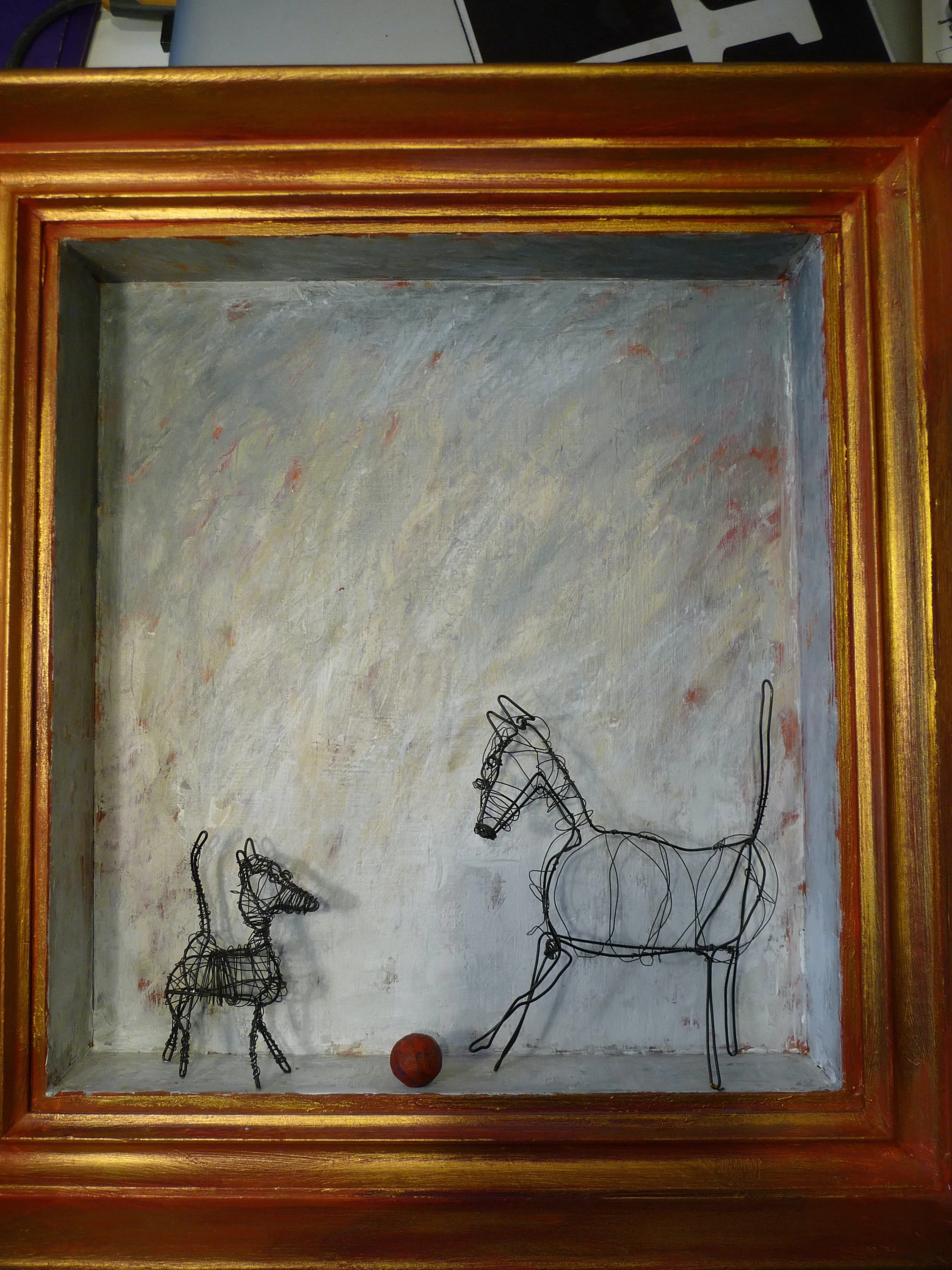 Två små statyer av wire, en hund och en häst, med en liten boll mellan dem, placerade i ett inramat fönster eller skåp.