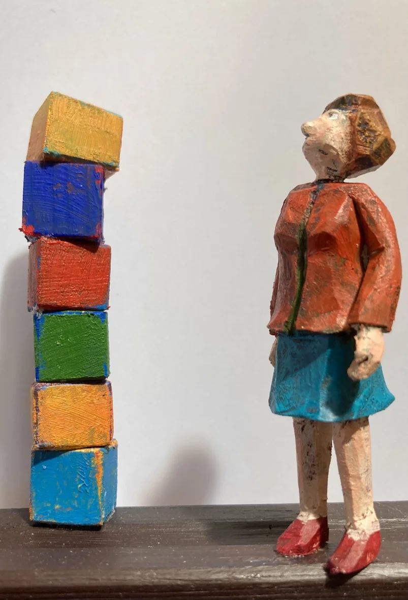 En liten träfigur av en flicka står bredvid några färgglada byggklossar.