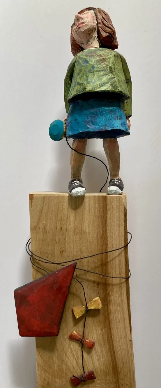 En konstnärlig skulptur av ett barn som håller en blå megafon, står på en träkloss med tråd och abstrakta former. Barnet har på sig gröna och blå kläder, och skulpturen är gjord i trä och färg.
