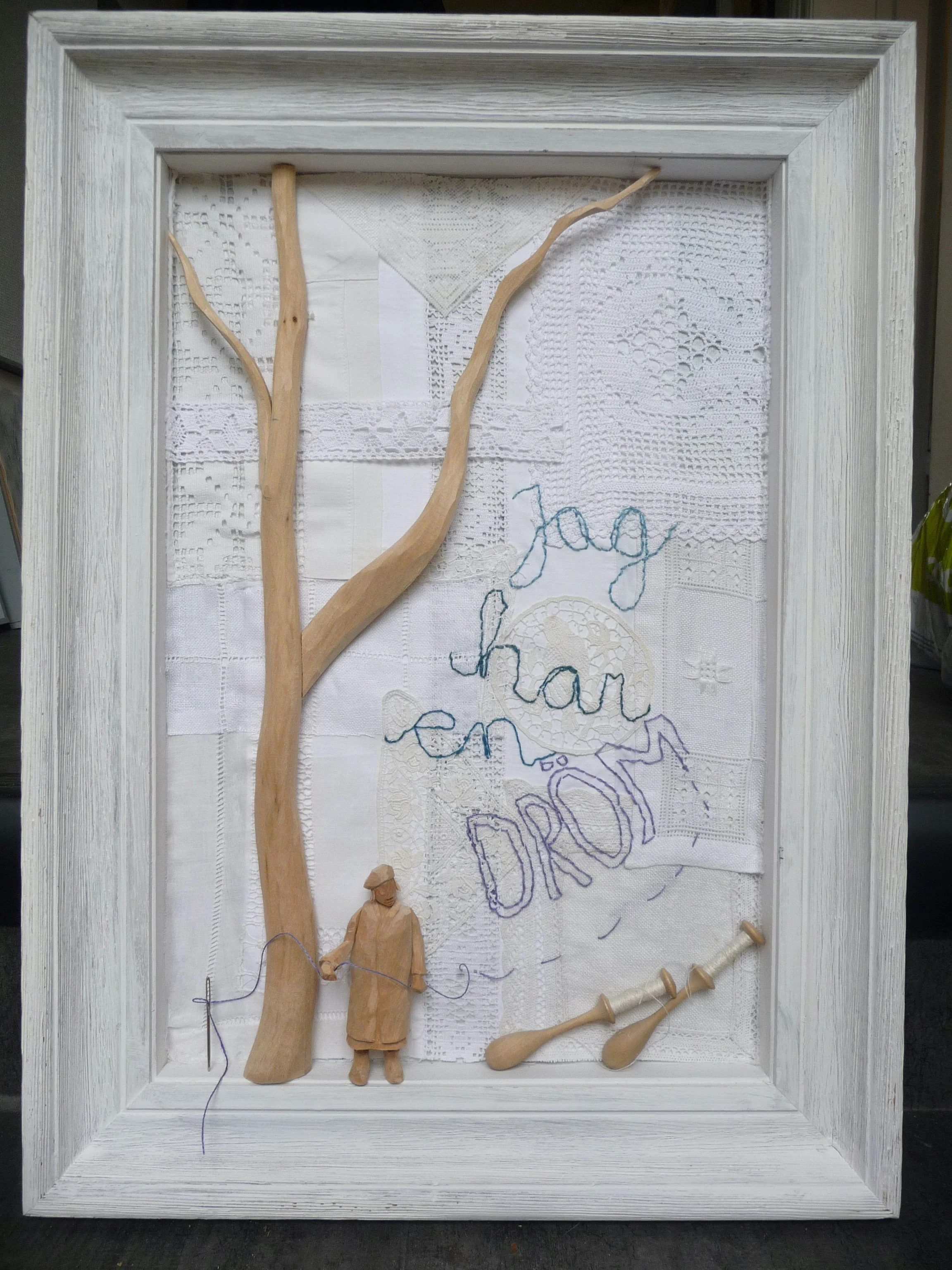 En vit träram med en väv av handgjorda vita lakan och tömda trädetjur och en liten trädocka, samt broderade ord 'jag har en dröm' i olika färger.