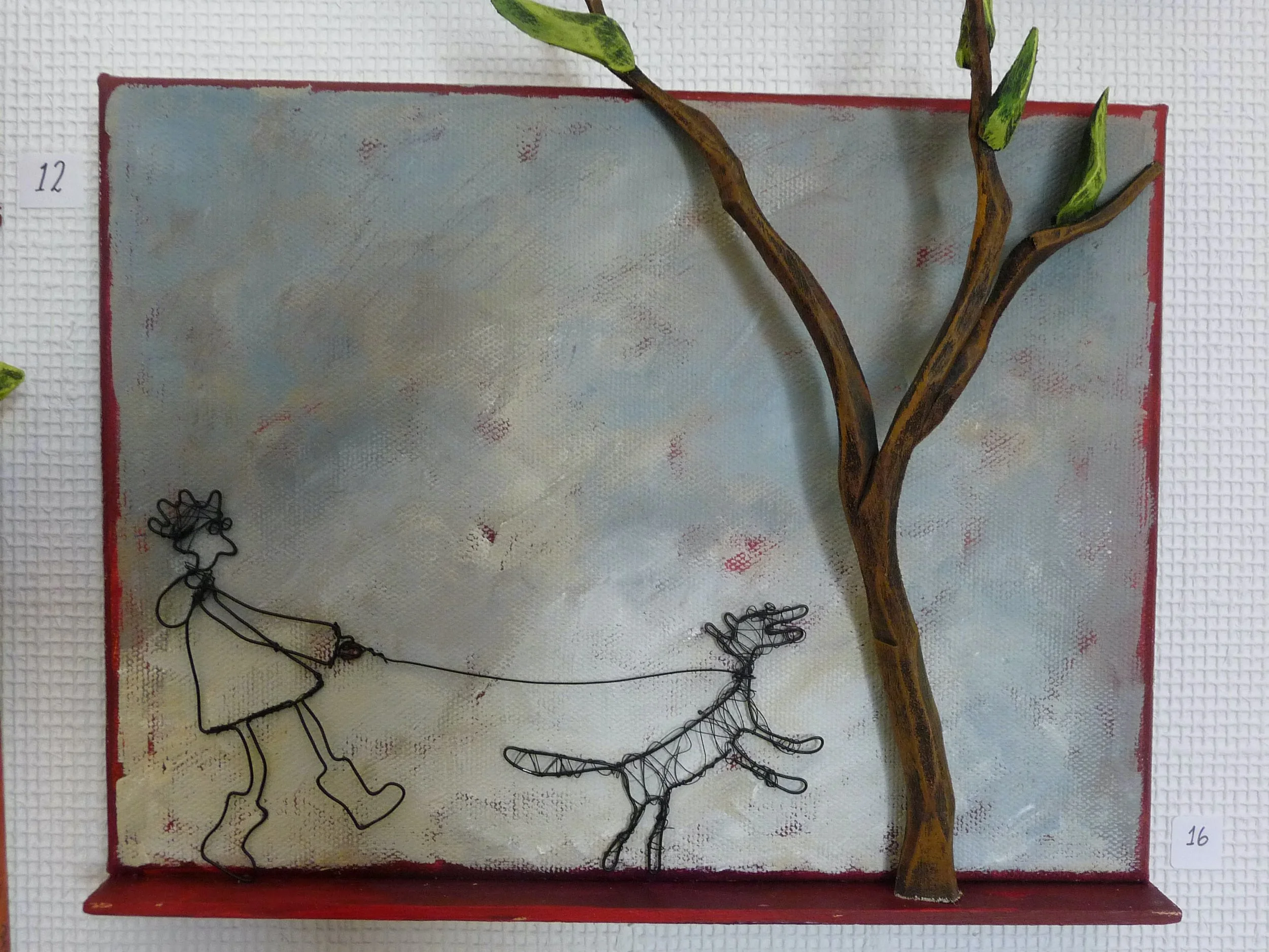 Konstverk med en person och en hund i wire, en trädgren och ett konstgjort träd, på en vit vägg.