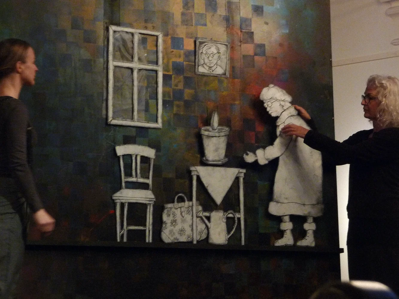 Två kvinnor interagerar med en väggmålning av en äldre kvinna som planterar en blomma i en blomkruka. Målningen föreställer en äldre kvinna i klänning och glasögon, som håller en blomma över ett bord, med en stol, en väskor och en kanna på golvet. De