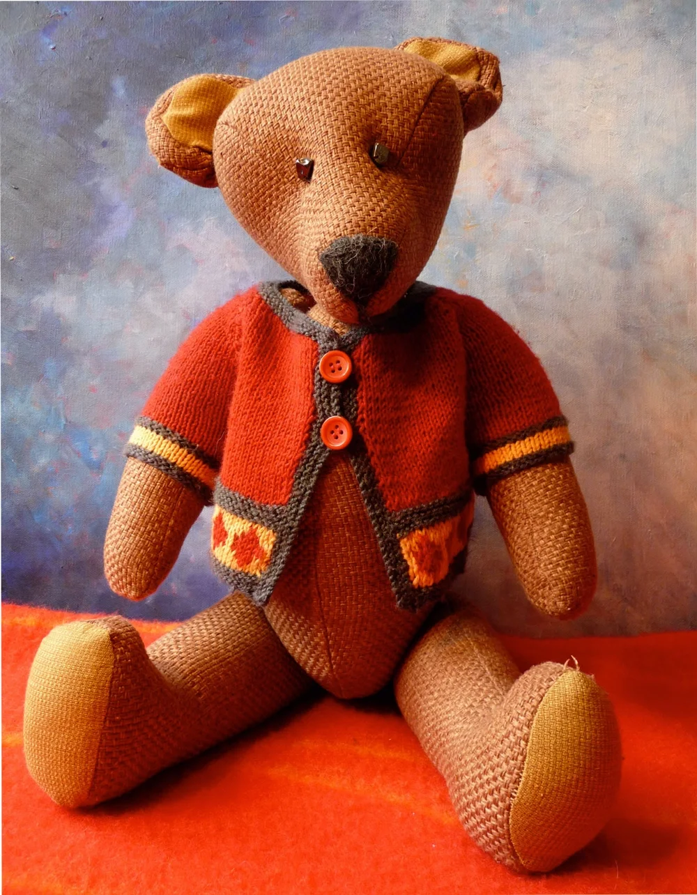 Såg en tygleksak i form av en nallebjörn med stickat utseende, klädd i en orange och grå tröja, sitthär i en sittposition på orange bakgrund.