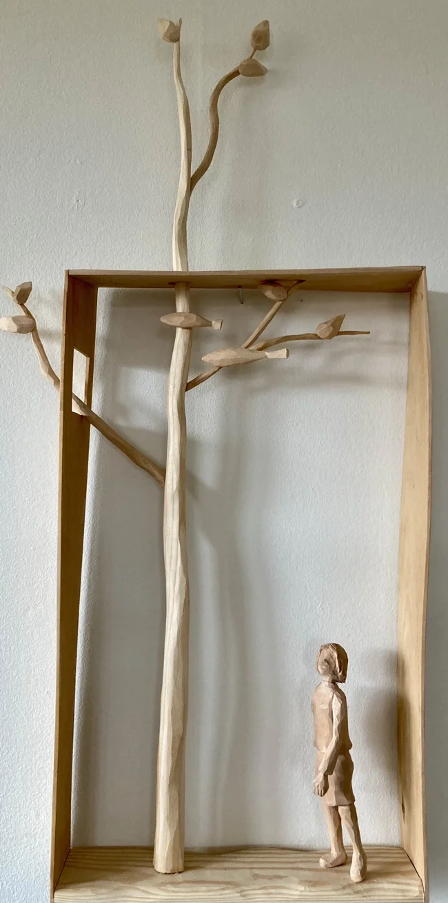 En träskulptur föreställande ett barn som står framför ett stort träd med knotiga grenar, inramad i en trälåda.