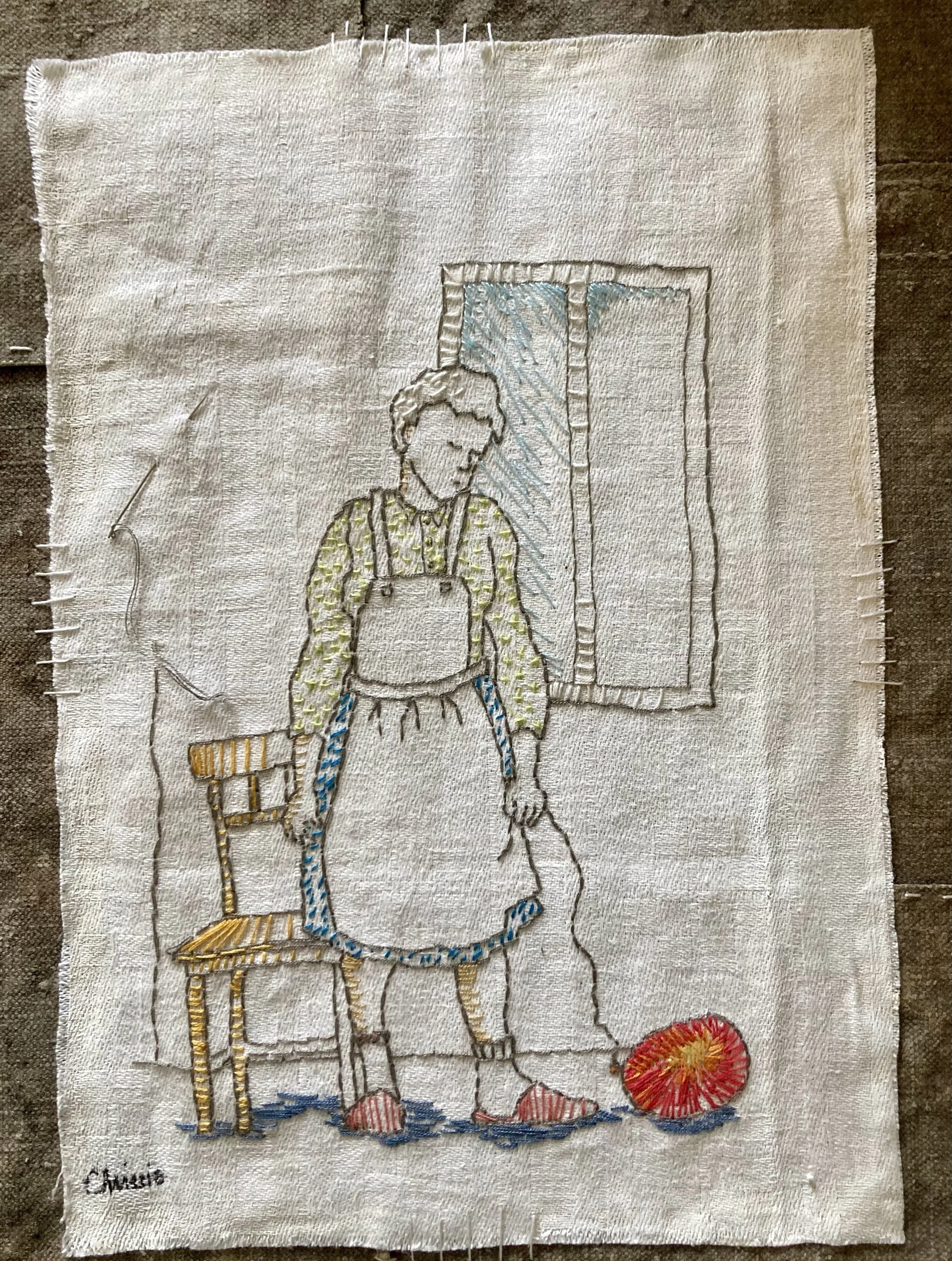 Handhållet broderi av en man i en skjorta och förkläde, stående vid en stol, med ett fönster i bakgrunden och en boll av garn på golvet.