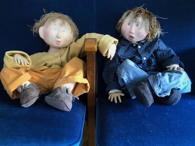 Två dockor sitter på en blå soffa, den vänstra klädd i gult och den högra i blått.