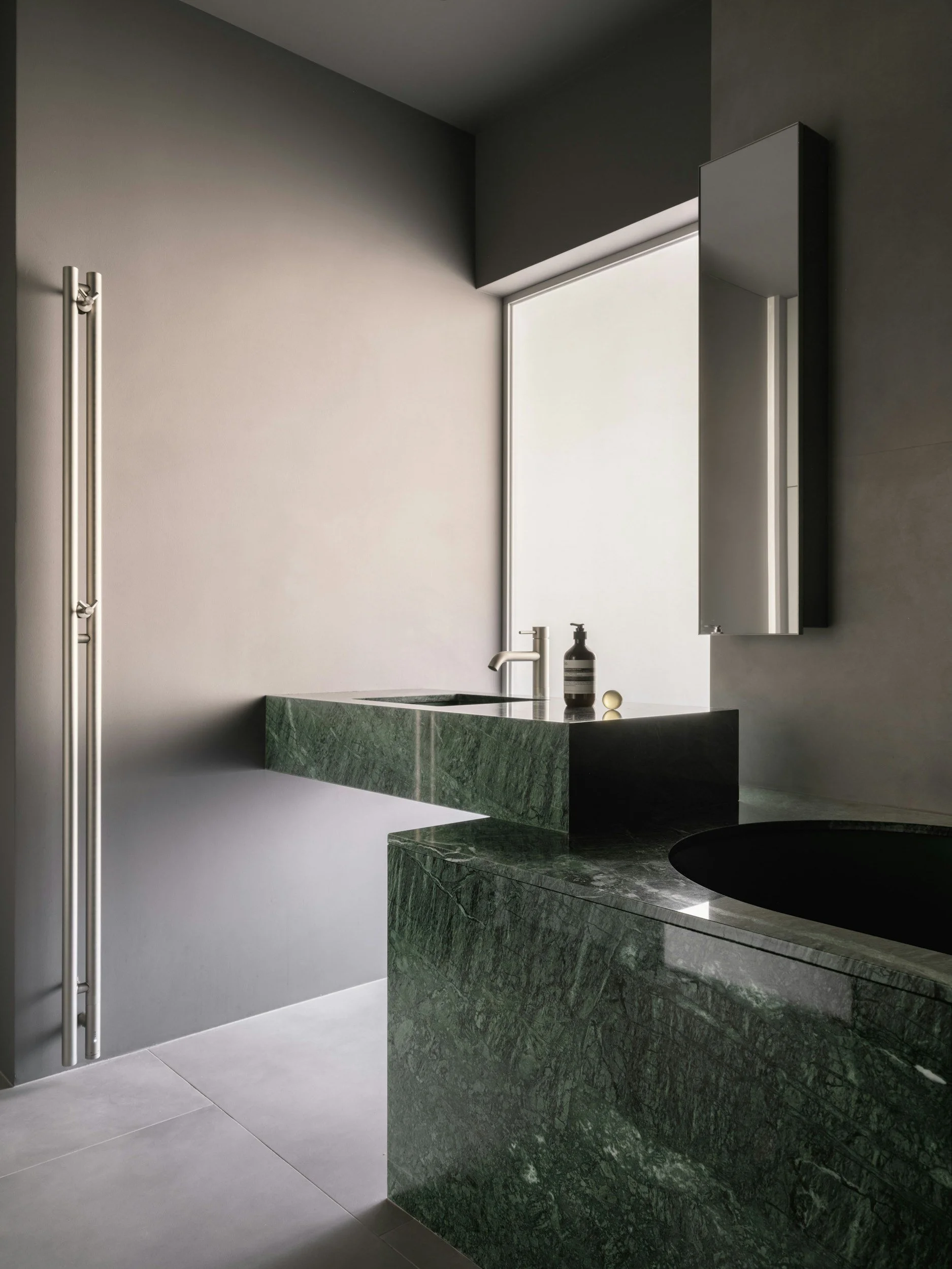 Modern bathroom with green marble sink and countertop, gray wall, mirror, soap dispenser, and towel radiator, with natural light from window.