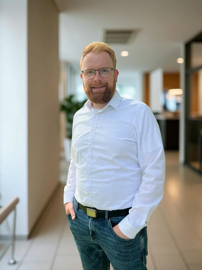Ein Mann mit Brille und rotem Haar, trägt ein weißes Hemd und blaue Jeans, steht in einem Innenraum mit heller Wand und moderner Einrichtung.