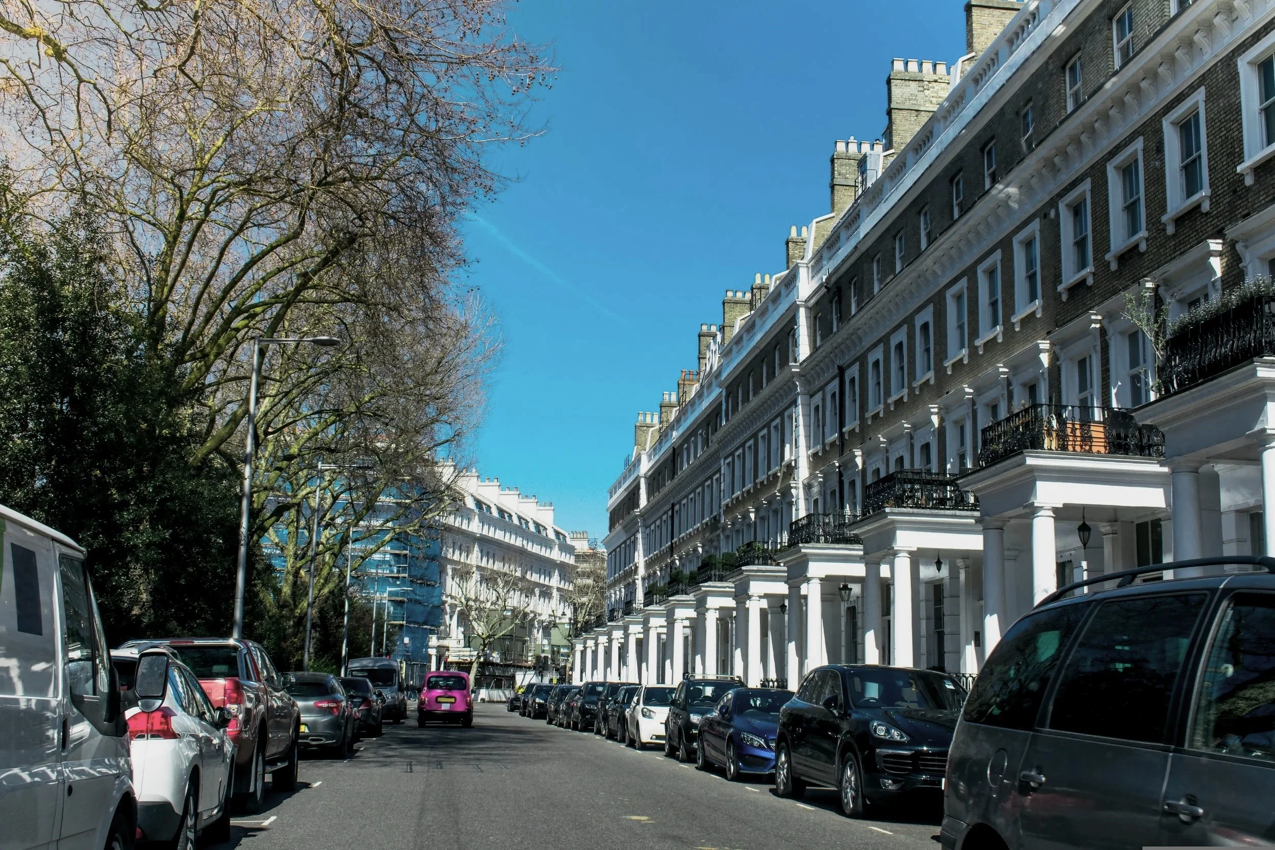 A street scene with parked cars and white buildings with black iron balconies on a sunny day.