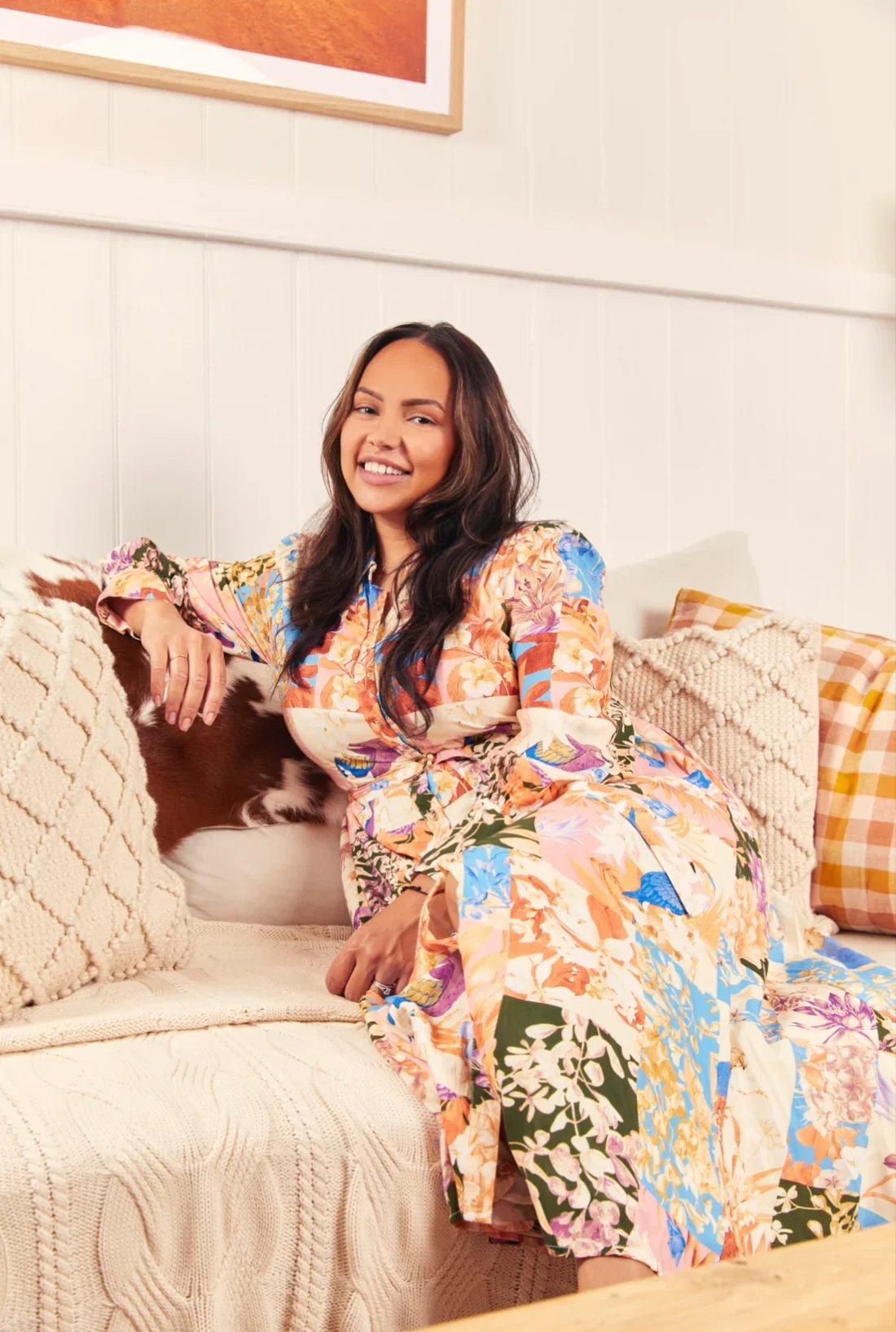 A woman with long dark hair sitting on a beige couch, smiling and wearing a colorful floral dress in a cozy, well-decorated living room.