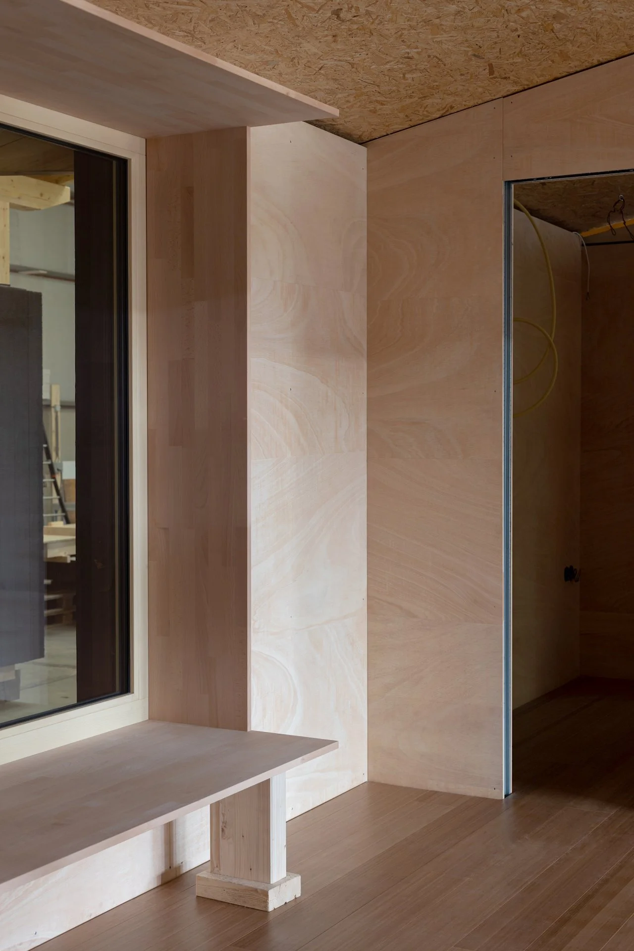Innenraum eines Gebäudes mit ungeputztem Holz und Baustoffen, inklusive Fenster mit schwarzem Rahmen und sichtbaren Kabeln, im Bau befindlich.