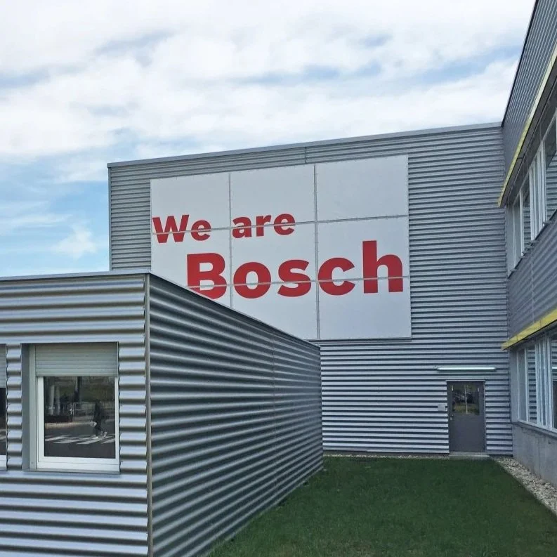 Außenansicht eines Gebäudes mit einem großen weißen Schild, auf dem steht 'We are Bosch' in roter Schrift.