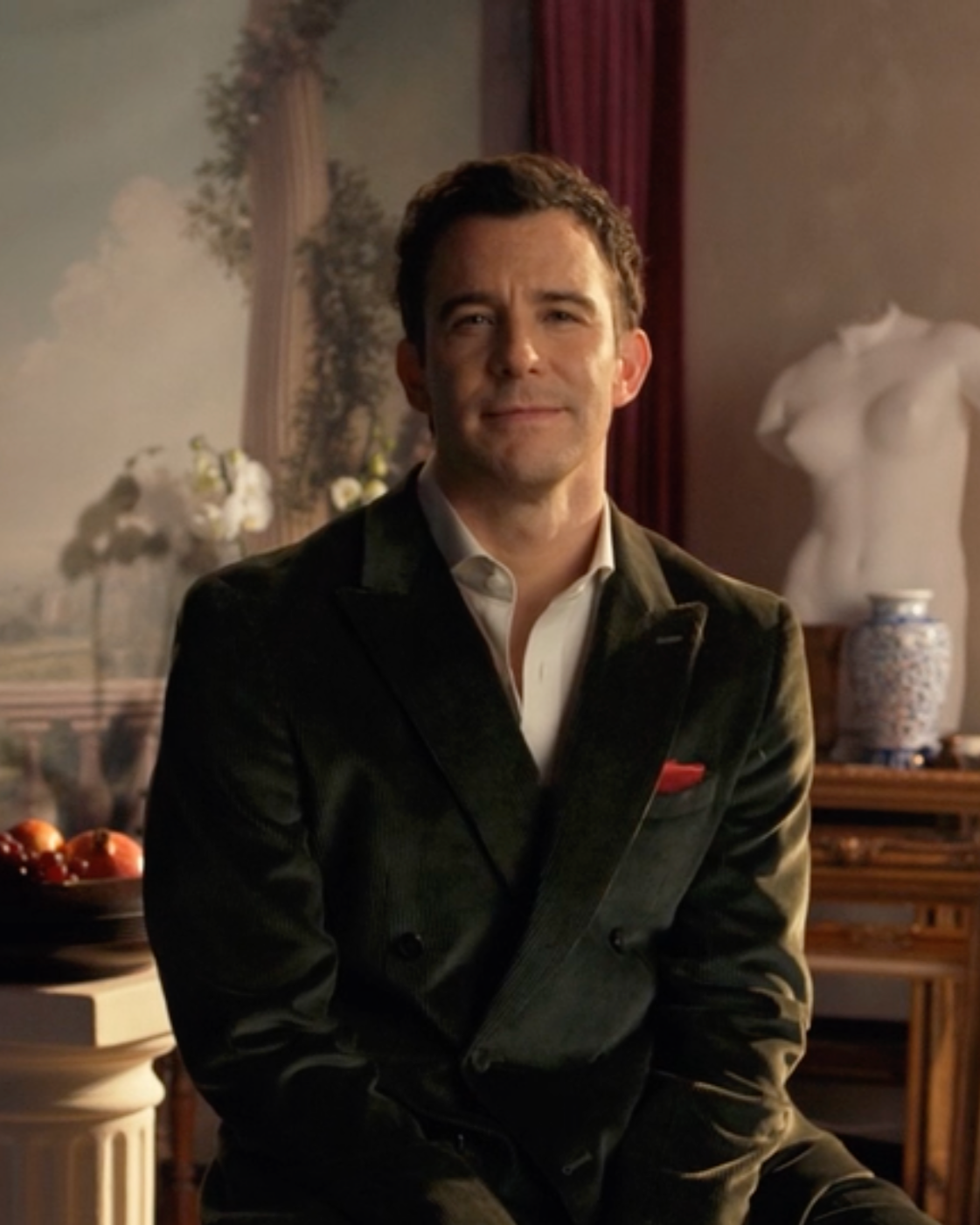 A man with dark hair and light skin, wearing a dark blazer and white shirt, sitting in a room with classical decor, including a female torso sculpture, a vase, and wall art of a landscape.