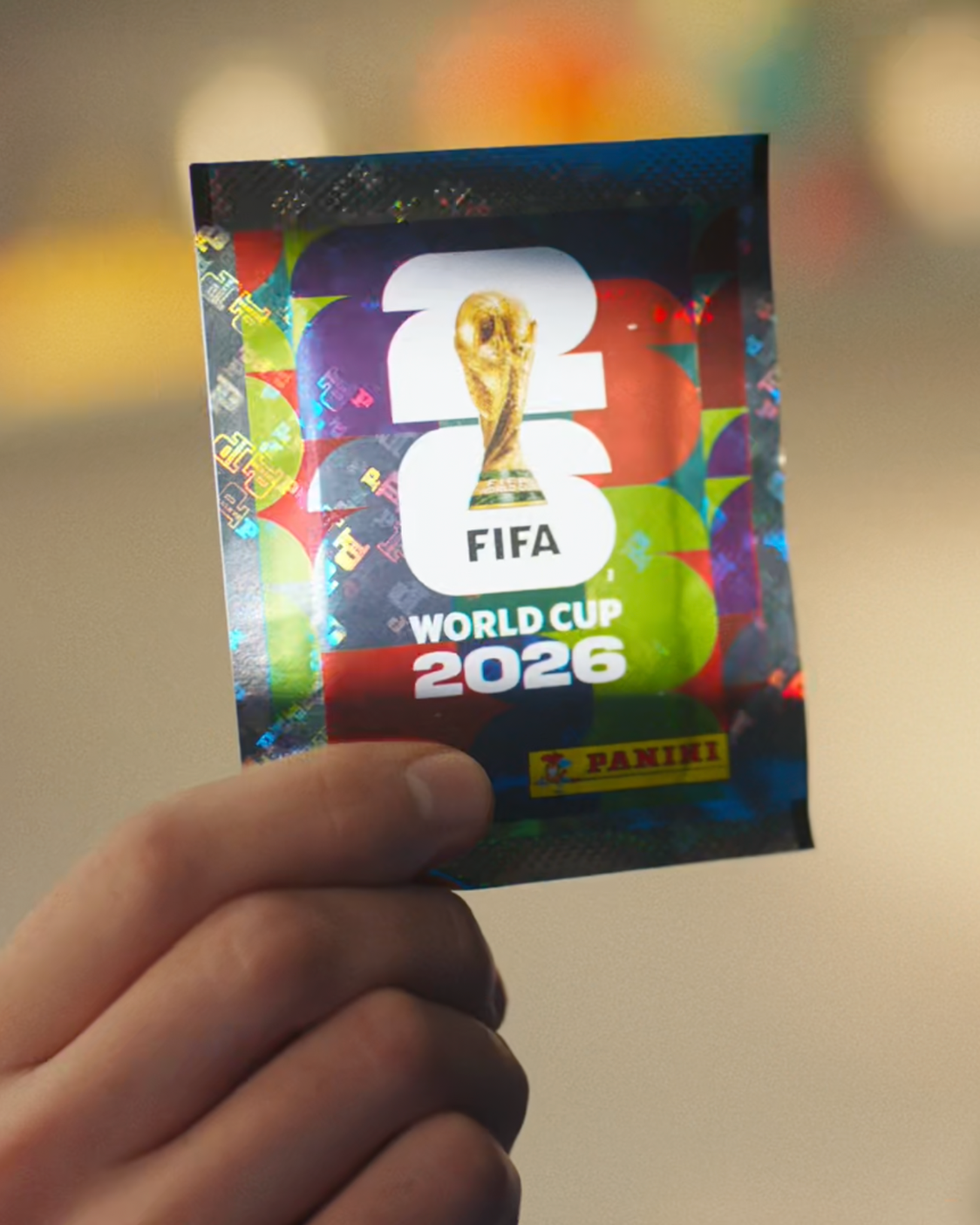 A hand holds a shiny, holographic Panini FIFA World Cup 2026 trading card featuring the FIFA World Cup trophy and colorful background.