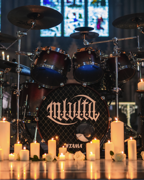 A drum set with the band name Melvins on the bass drum, surrounded by lit candles and flowers in a church or cathedral setting.