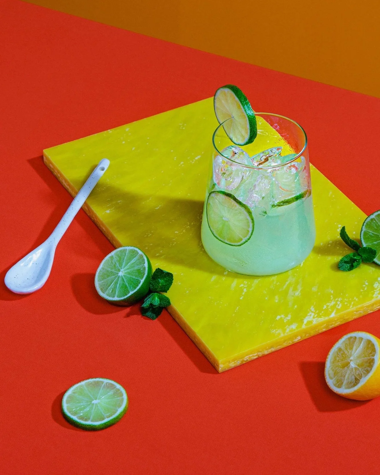 Glass of lemonade with lime slices and ice on yellow and orange background with lemon halves and lime slices scattered around.