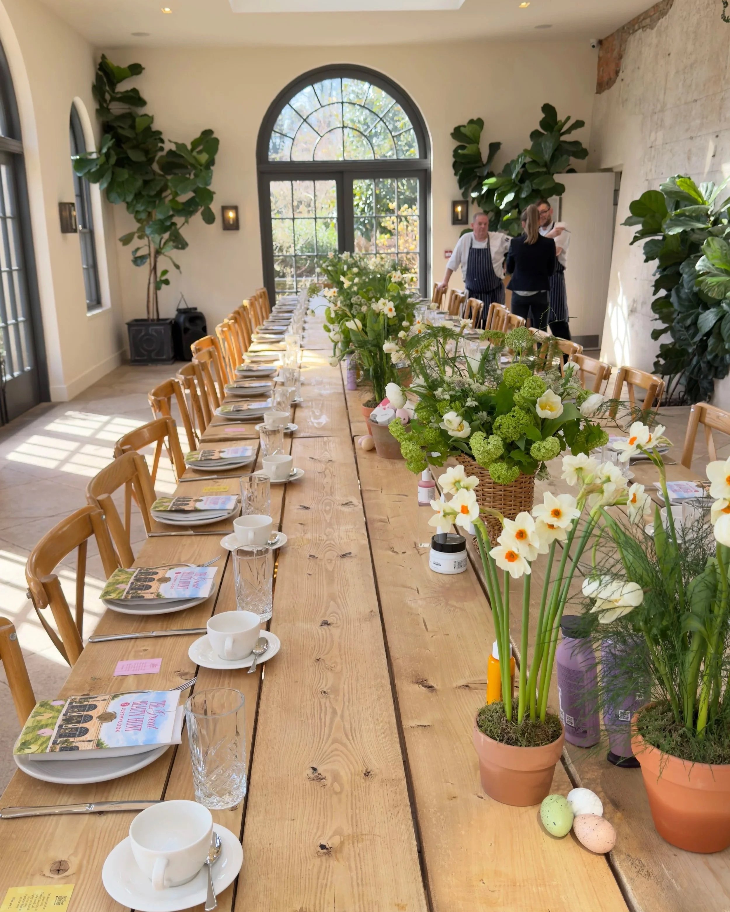 Long wooden dining table set for a meal with white plates, cups, glasses, and cutlery, decorated with large potted plants with vibrant flowers, in a bright room with large arched window and people in kitchen attire and formal clothing talking at the far end.
