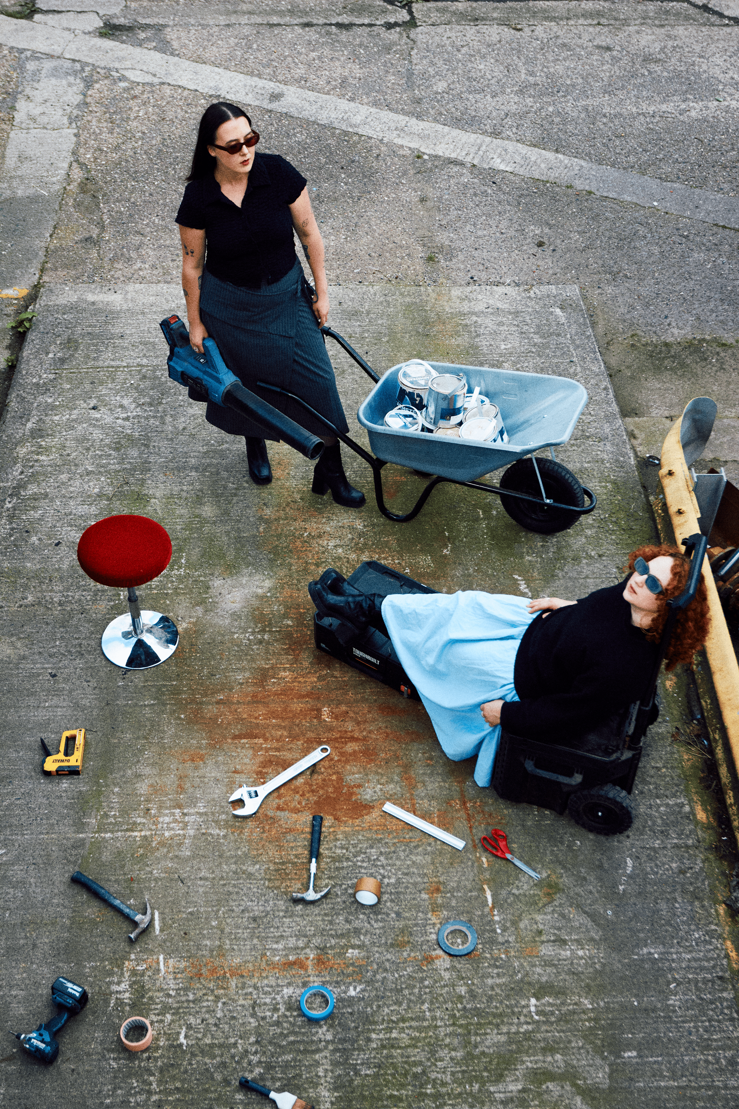 An overhead view of two women, one standing and one lying on a black scooter, surrounded by scattered tools on a weathered concrete surface. The standing woman wears sunglasses, a black shirt, and a long skirt, holding a leaf blower. The woman on the scooter has curly red hair, sunglasses, and is wearing a black top and a light blue skirt.