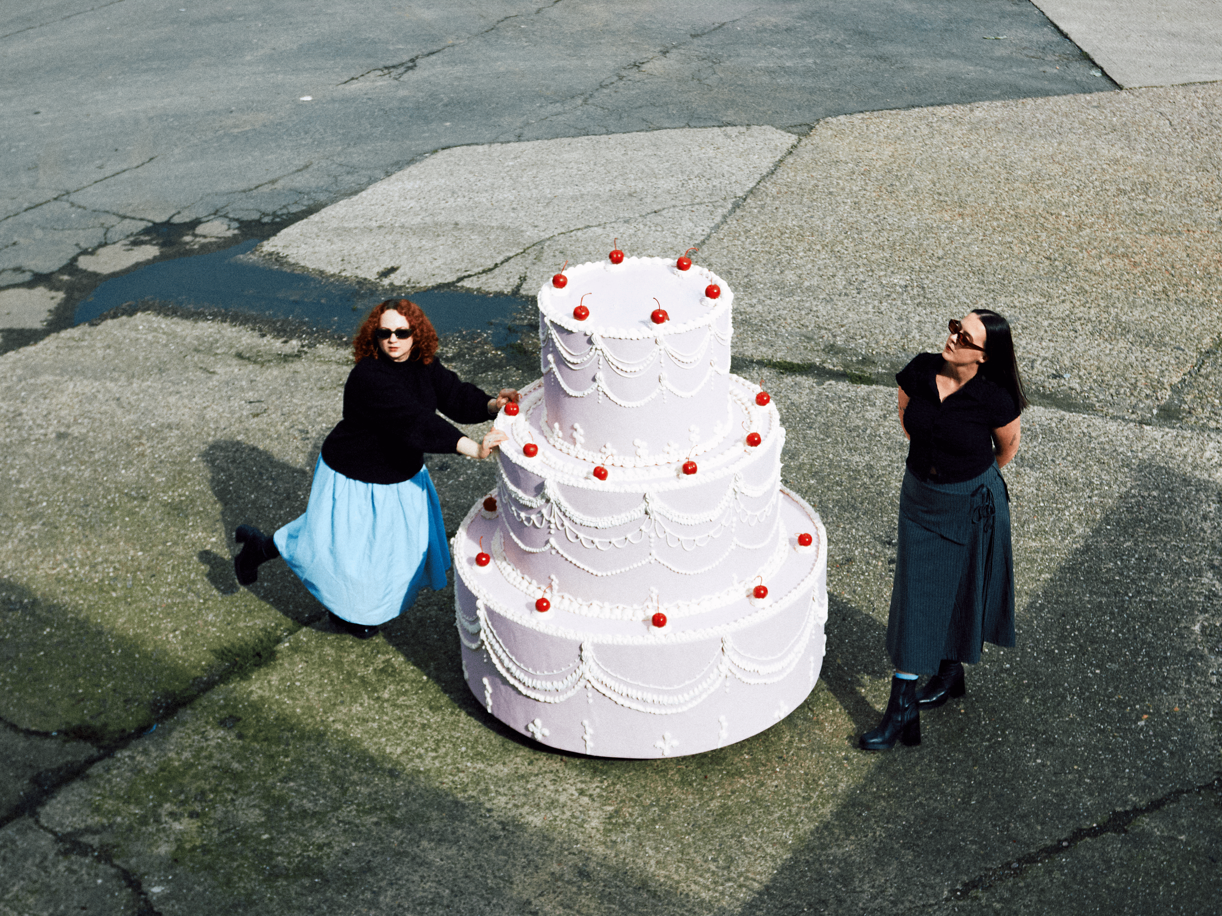 Two women are standing beside a multi-tiered white cake decorated with red cherries and white piped icing, on an outdoor concrete surface.