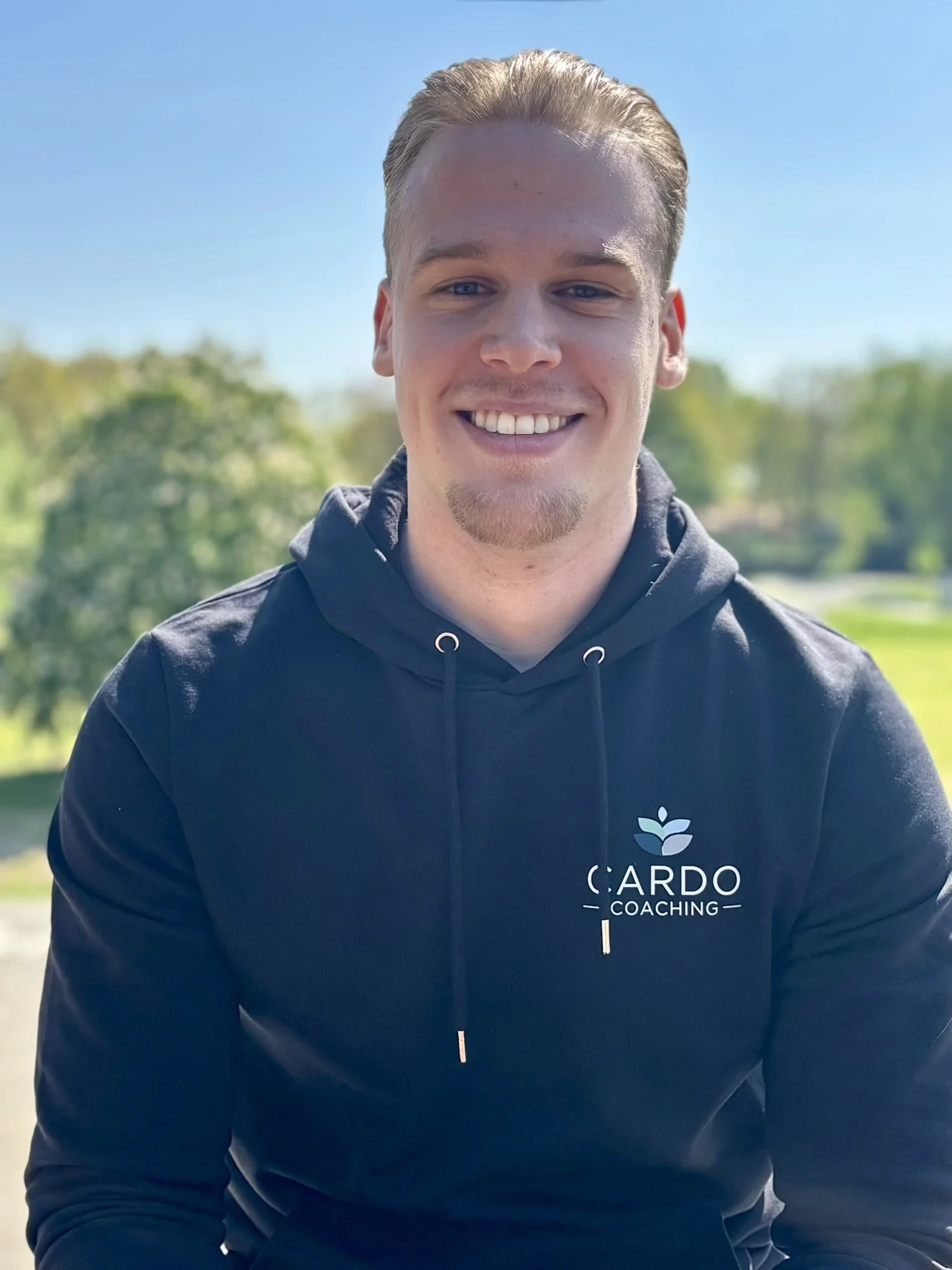 Close-up van een glimlachende man in een zwart hoodie met het logo 'CARD COACHING' op de voorkant, buiten op een zonnige dag met groene bomen op de achtergrond.