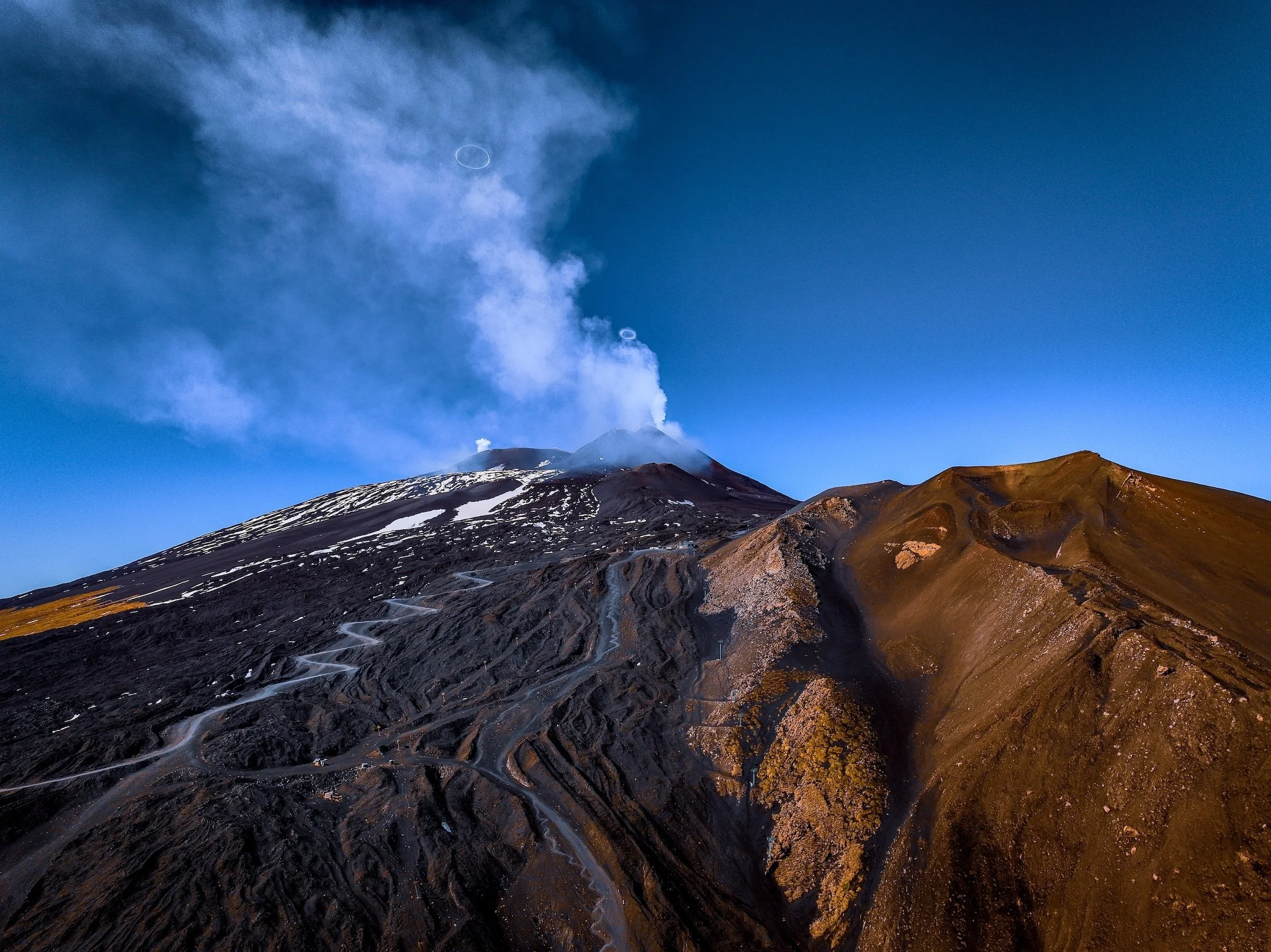 Vulkan mit Rauch und Asche, graue Lavaflüsse auf der schwarzen Vulkanoberfläche, umgeben von braunem, trockenem Gelände, blauer Himmel im Hintergrund.