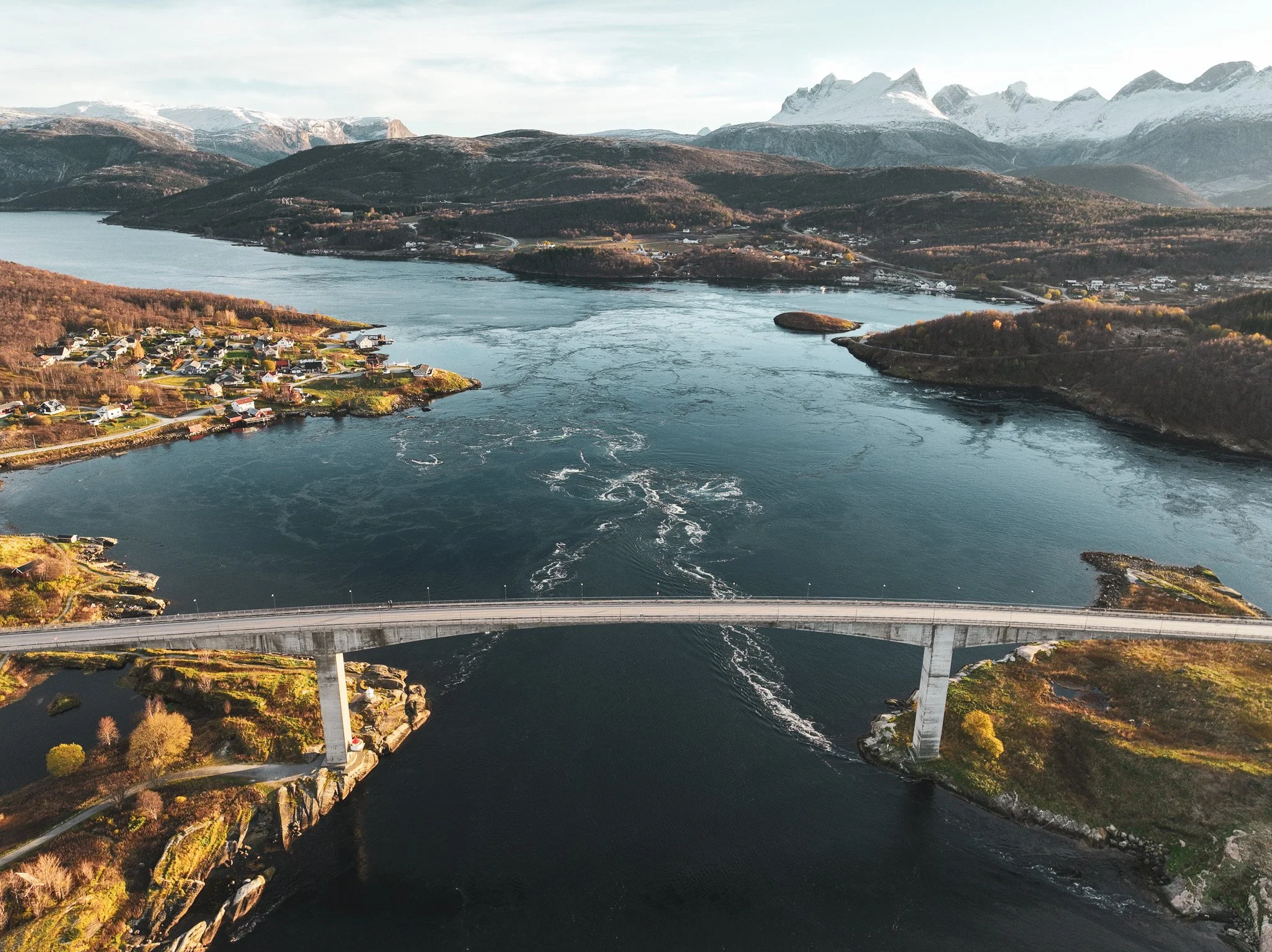 Luftaufnahme einer Brücke, die über einen Fluss führt, mit kleinen Häusern am Flussufer und Bergen im Hintergrund.
