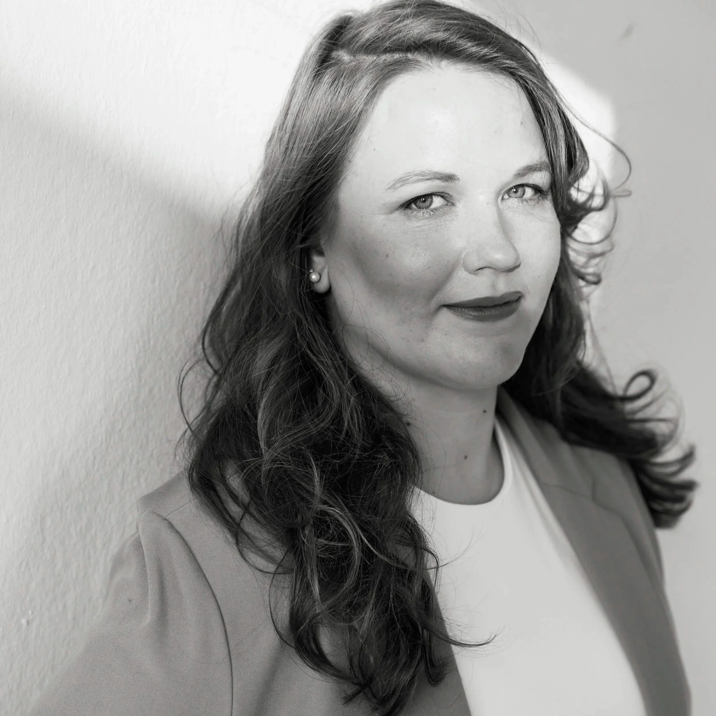 Schwarz-Weiß-Porträt einer Frau mit langen, gewellten Haaren und einem leichten Lächeln, stehend an einer Wand.